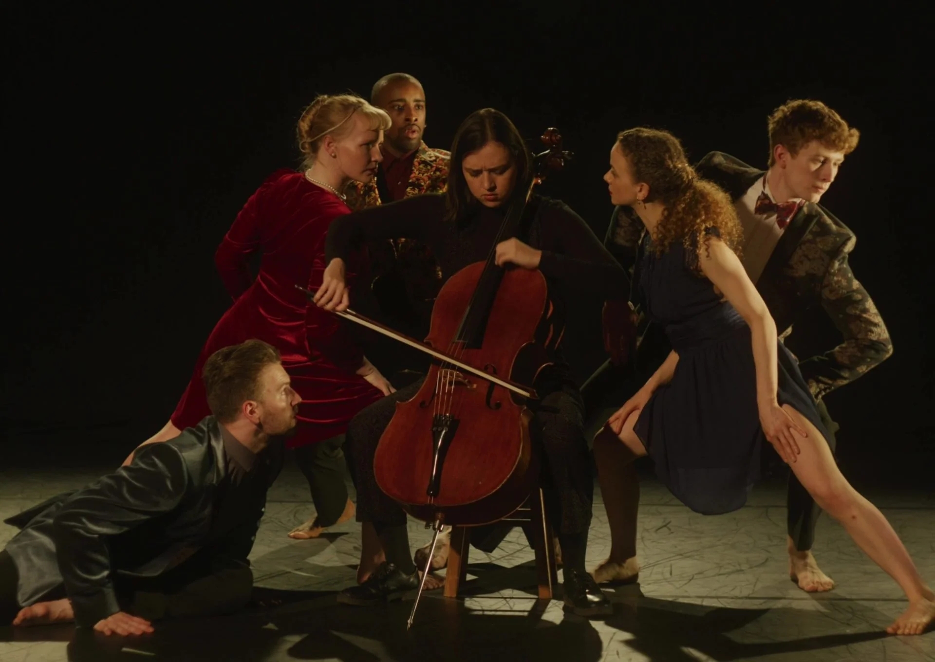 Group of six performers on stage, some kneeling or sitting, surrounding a woman playing a cello, with intense expressions, set against a dark background.