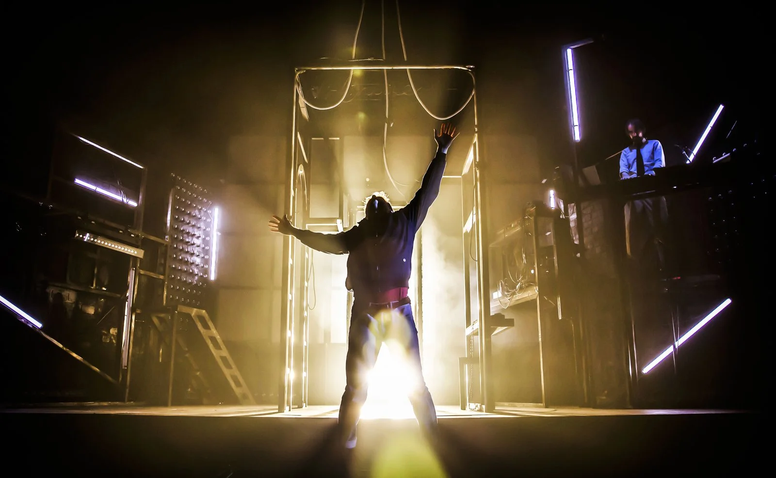 Person with arms raised standing in front of glowing light on stage, with a person at a DJ booth illuminated by neon lights to the side.
