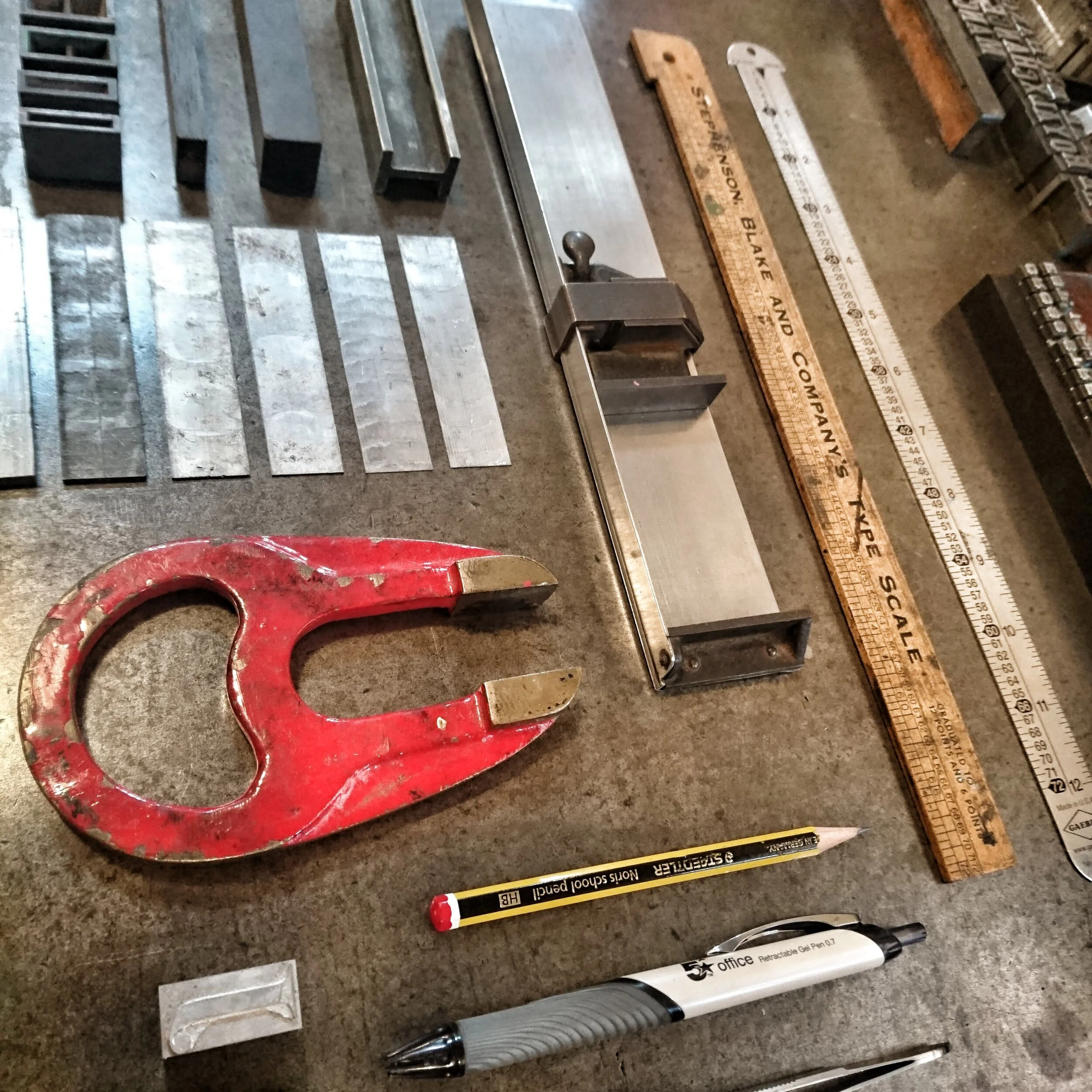 Various Letterpress tools and materials on a workbench, including letterpress typescales, leads, composing sticks, a pencil, a pen, and a and letterpress furniture