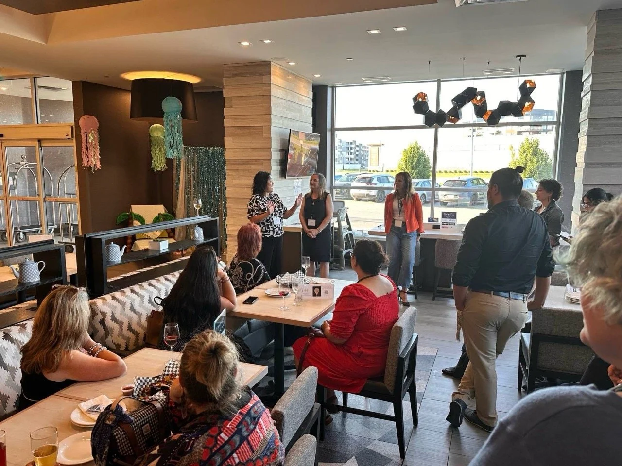 A group of women standing and seated in a modern restaurant or cafe, attending a presentation or speech with three women speaking at the front near a large window with tables and chairs around.