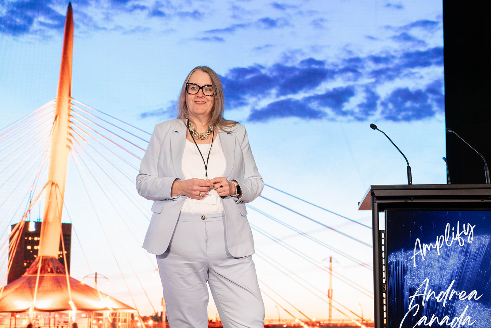 Andrea Canada standing on stage with a large background image of a bridge and a partly cloudy sky. 