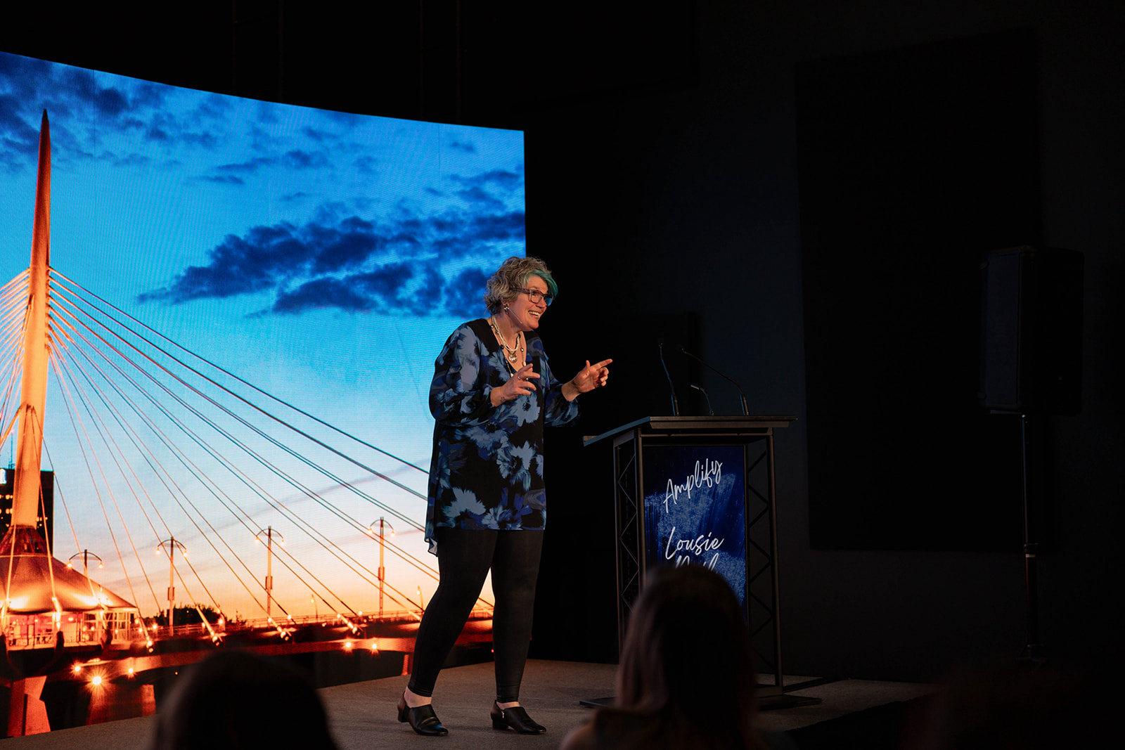 Louise Neil speaking on stage at Amplify conference with large screen in background displaying a bridge and clouds during sunset.