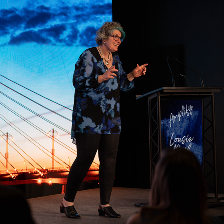 Louise Neil speaking on stage at Amplify conference with large screen in background displaying a bridge and clouds during sunset.