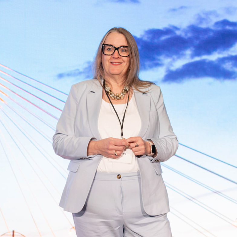 Andrea Canada standing on stage with a large background image of a bridge and a partly cloudy sky. 