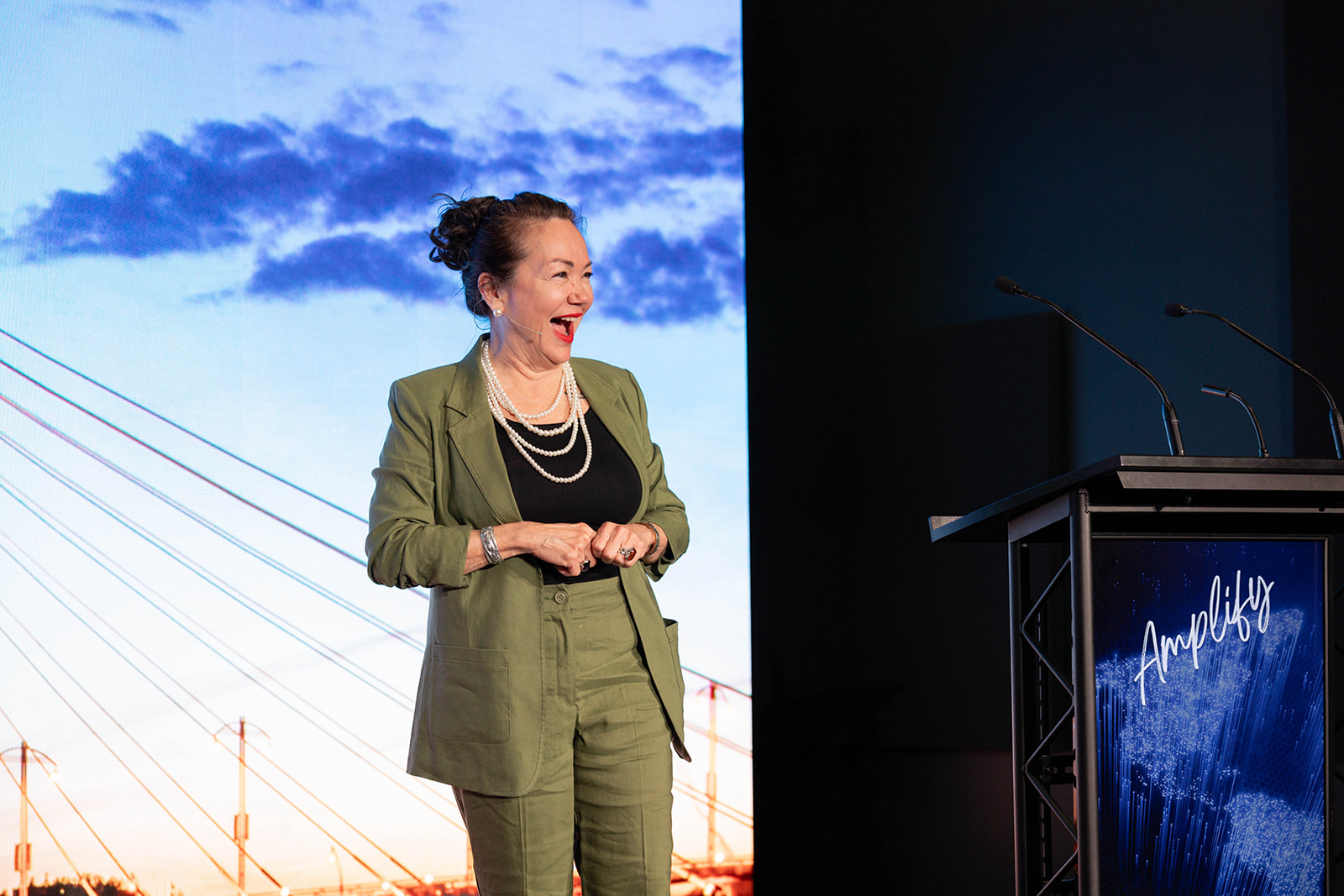 Denise Harder speaking on stage at Amplify, smiling and laughing, with a backdrop of a sky with clouds and a bridge. 