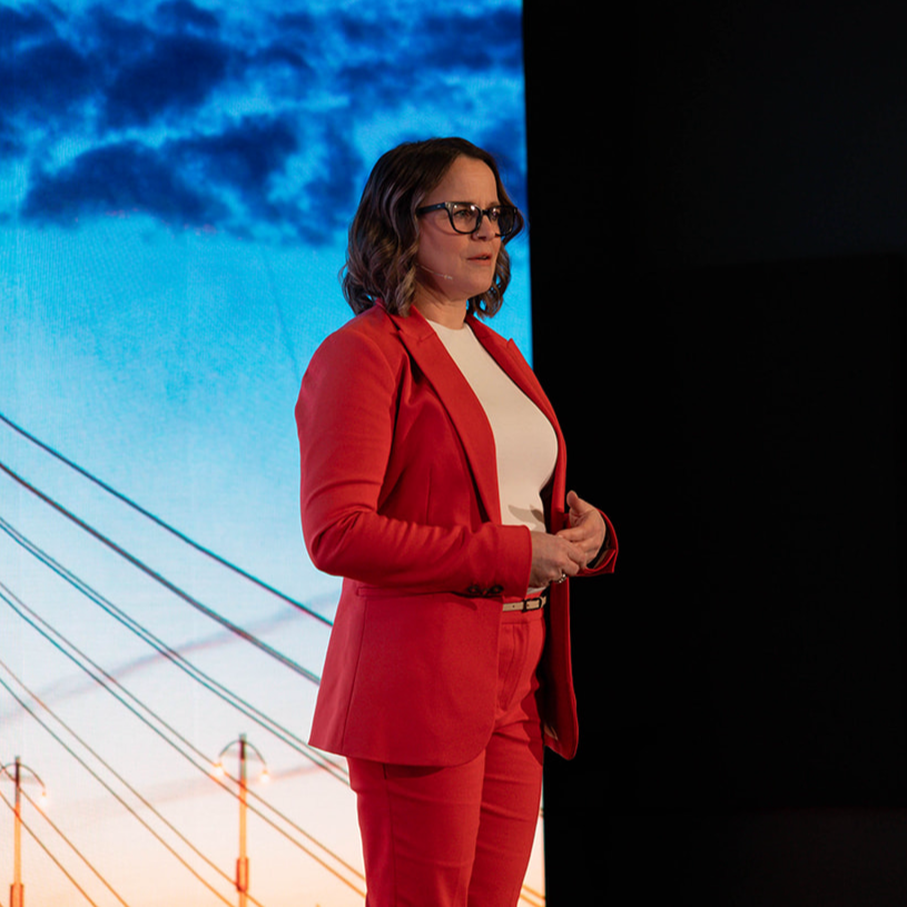 Kim Mintenko standing on stage with a presentation screen behind her displaying a bridge with a blue sky and clouds.