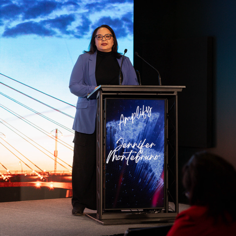 Jennifer Montebruno standing at a podium giving a presentation at Amplify with a large screen behind her displaying a sky with clouds and a bridge. 