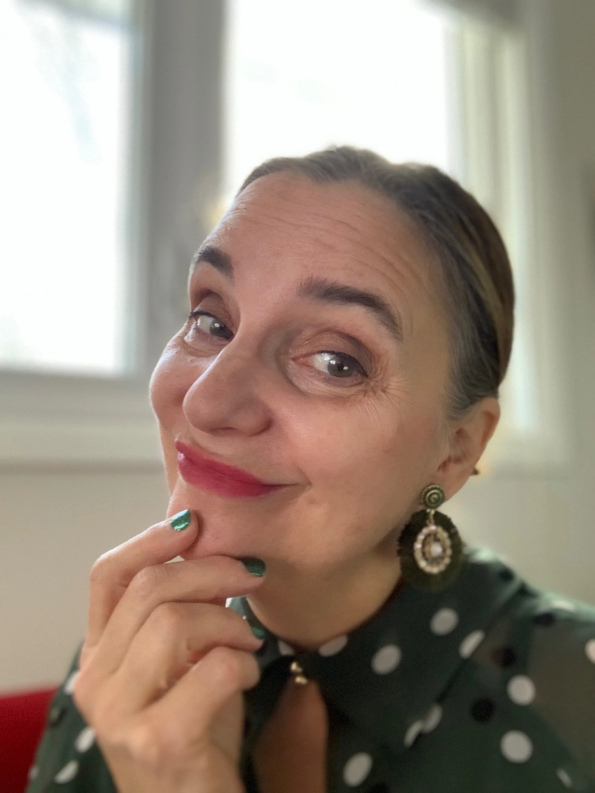 A woman with short dark hair styled back, wearing a polka dot blouse, red lipstick, and ornate earrings, posing with her hand near her chin and winking at the camera.