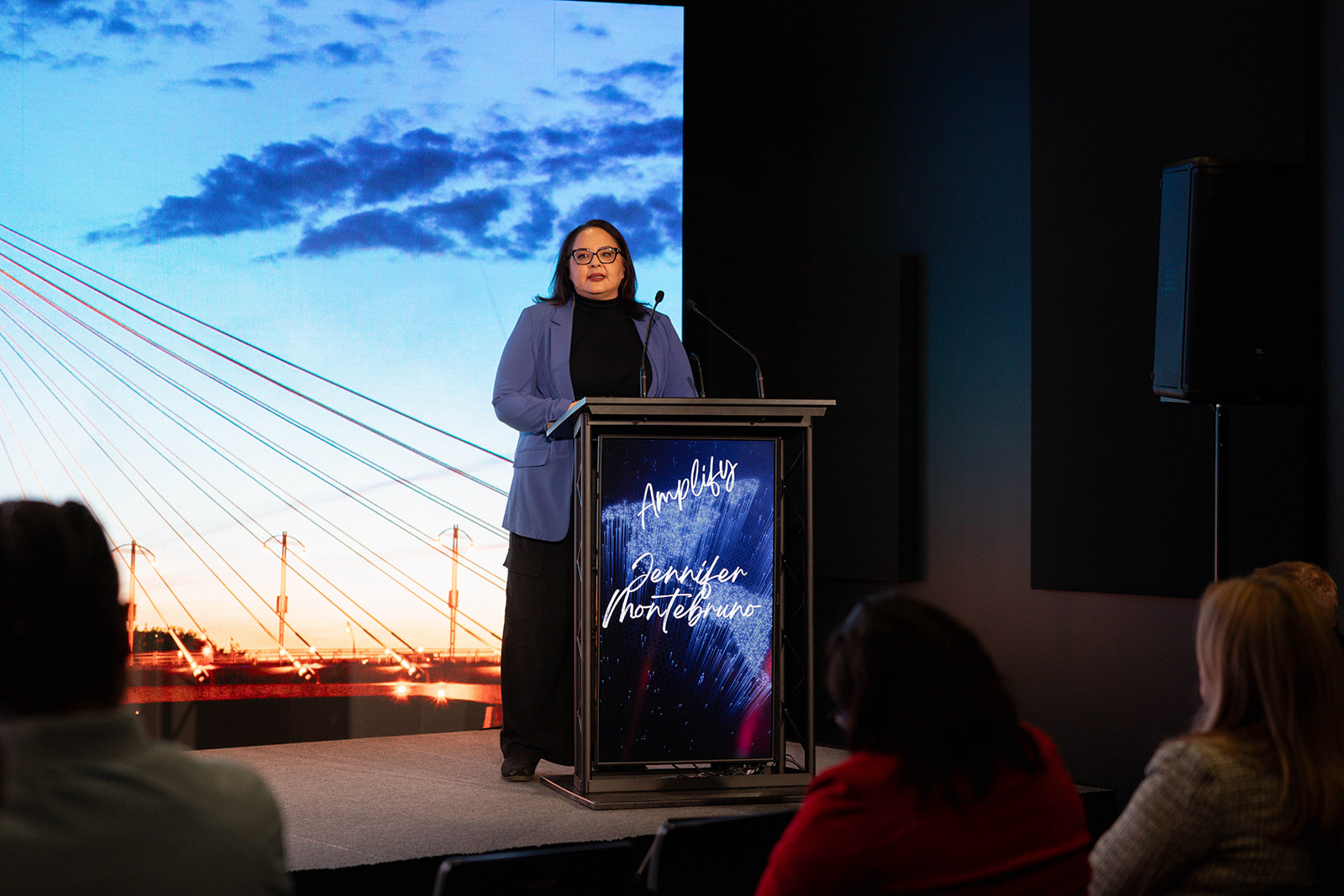 Jennifer Montebruno standing at a podium giving a presentation at Amplify with a large screen behind her displaying a sky with clouds and a bridge. 