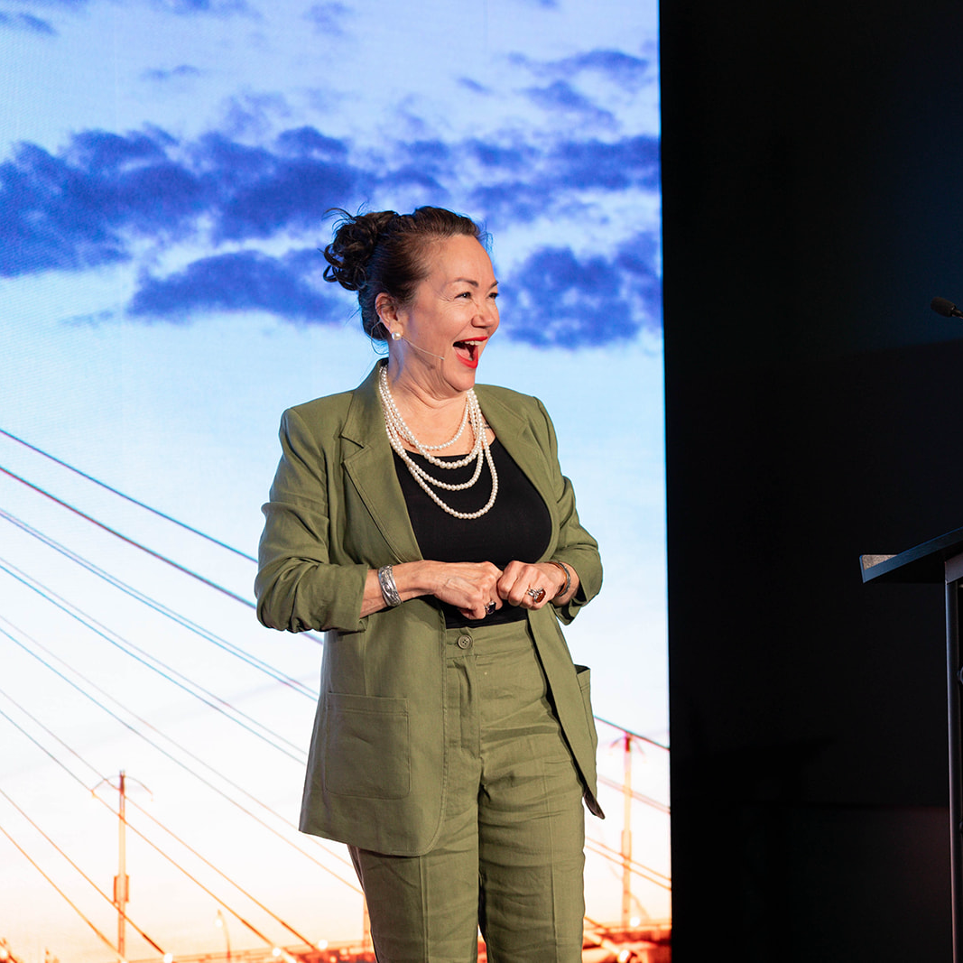 Denise Harder speaking on stage at Amplify, smiling and laughing, with a backdrop of a sky with clouds and a bridge. 