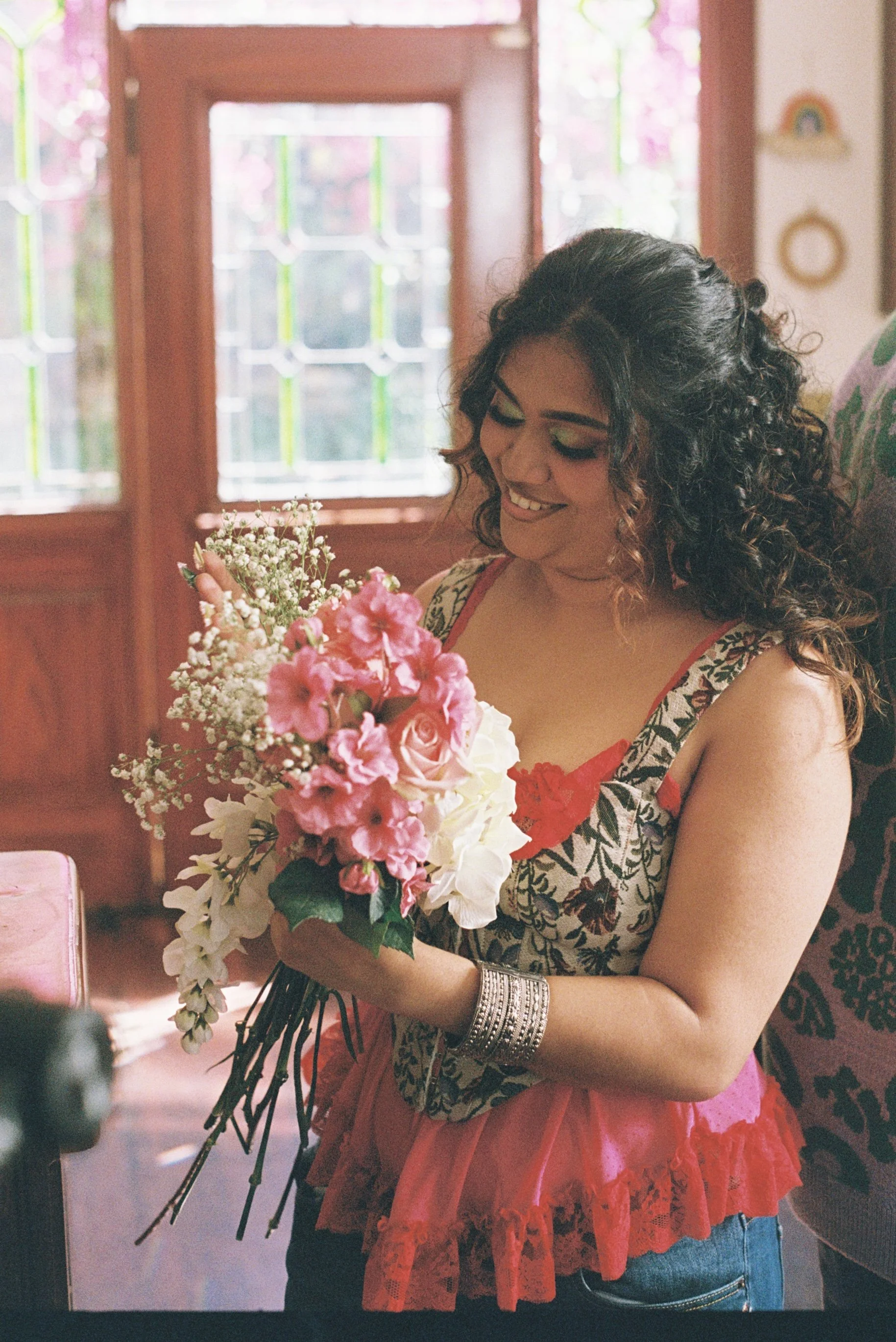 Woman with curly dark hair wearing a patterned sleeveless top and red lace skirt, smiling and holding a bouquet of pink and white flowers inside a room with stained glass windows.