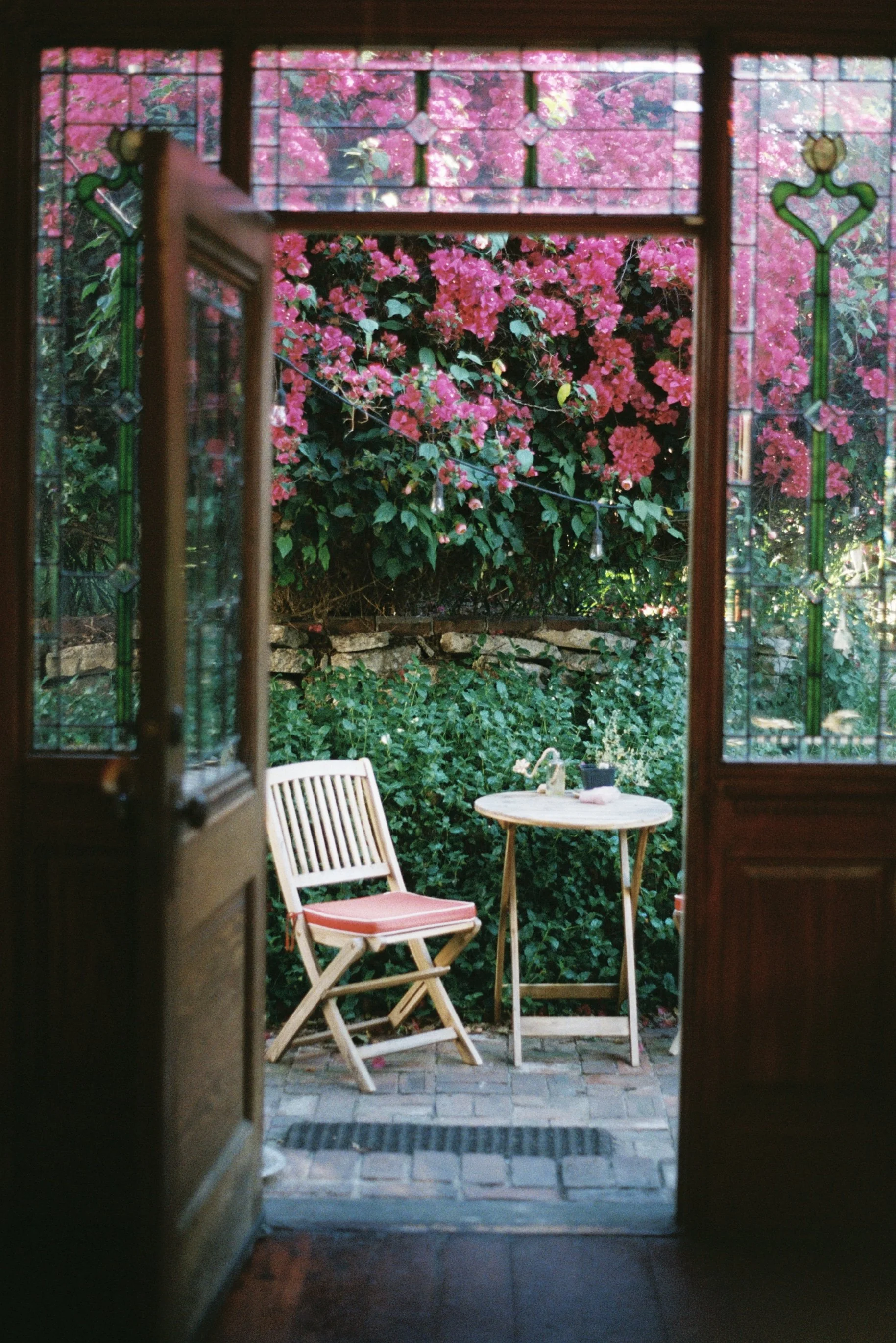 View through an open wooden door to a small outdoor patio with a round table, a folding chair with pink cushion, surrounded by pink bougainvillea flowering bushes and green foliage.