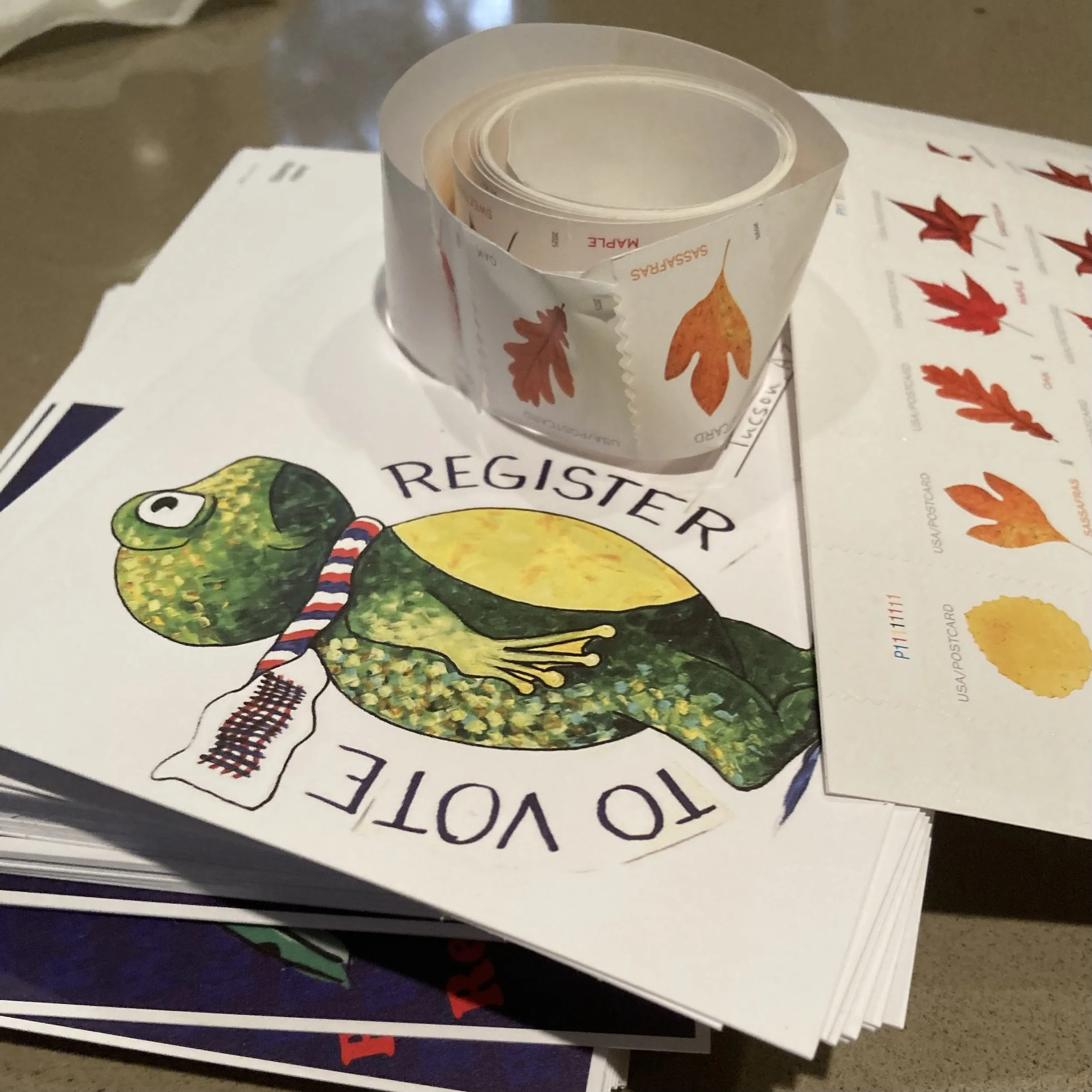A stack of postcards with a frog that says "Register to Vote", with a roll of postcard stamps on top.