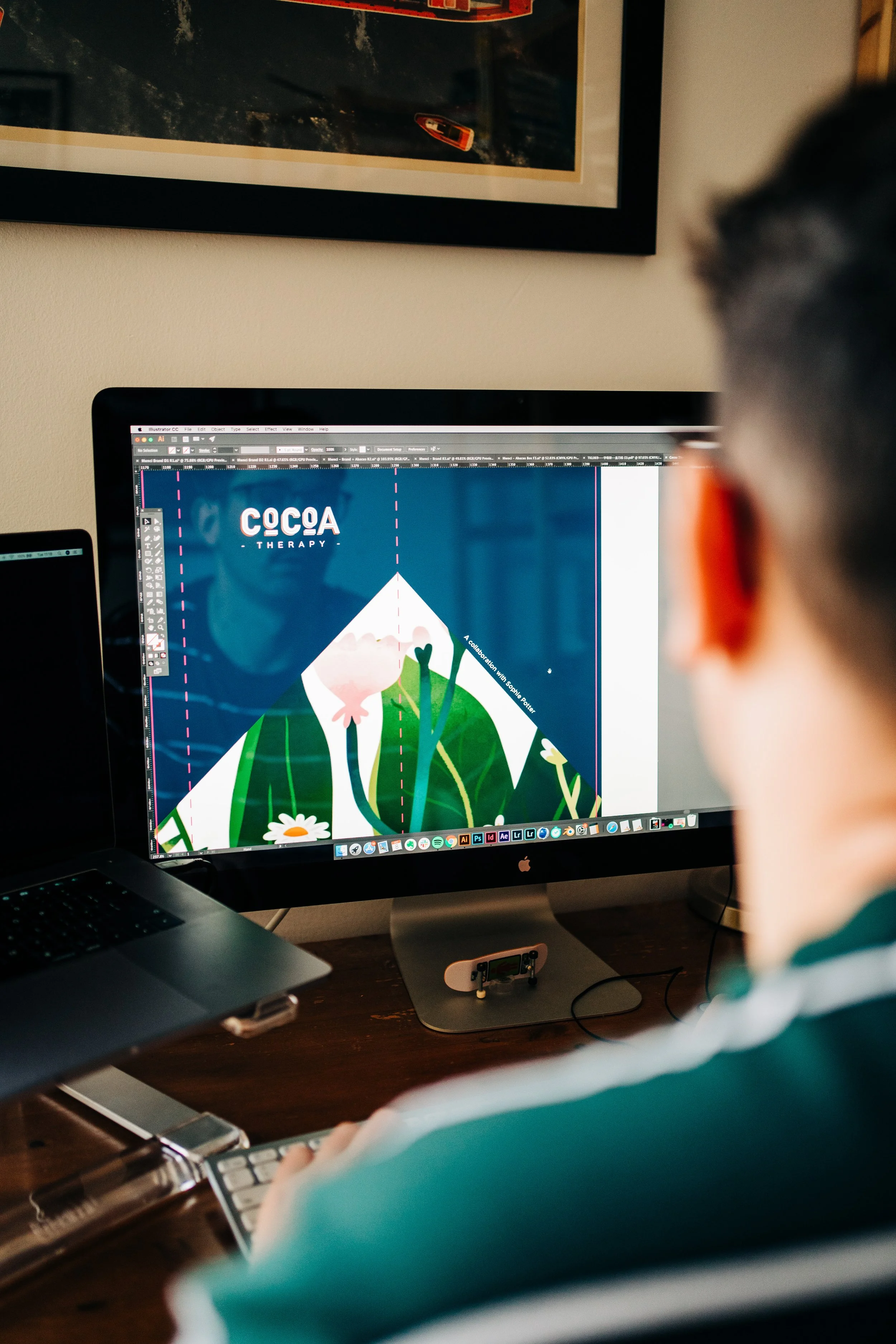 A person working on a creative digital design on an iMac computer, with an illustration featuring flowers and the text 'COCA THERAPY' on the screen.