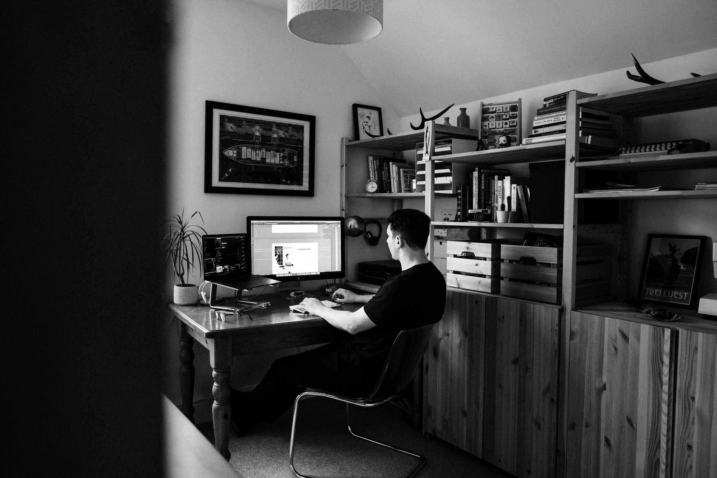 A person working at a desk in a home office, with multiple monitors, bookshelves, and framed pictures in a black-and-white photograph.
