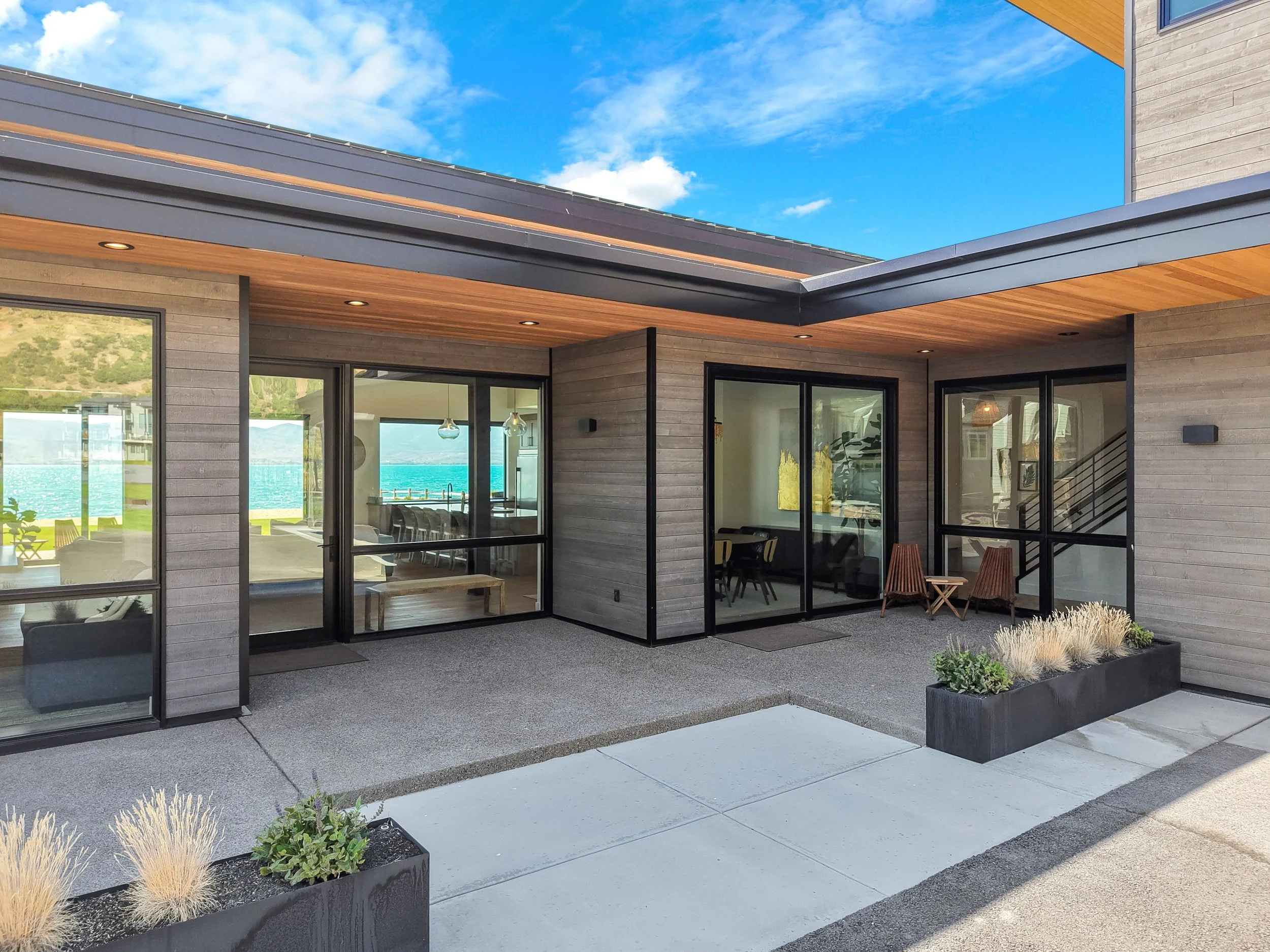 Modern house exterior with large glass sliding doors, wooden siding, and potted plants outside, overlooking water and hills.