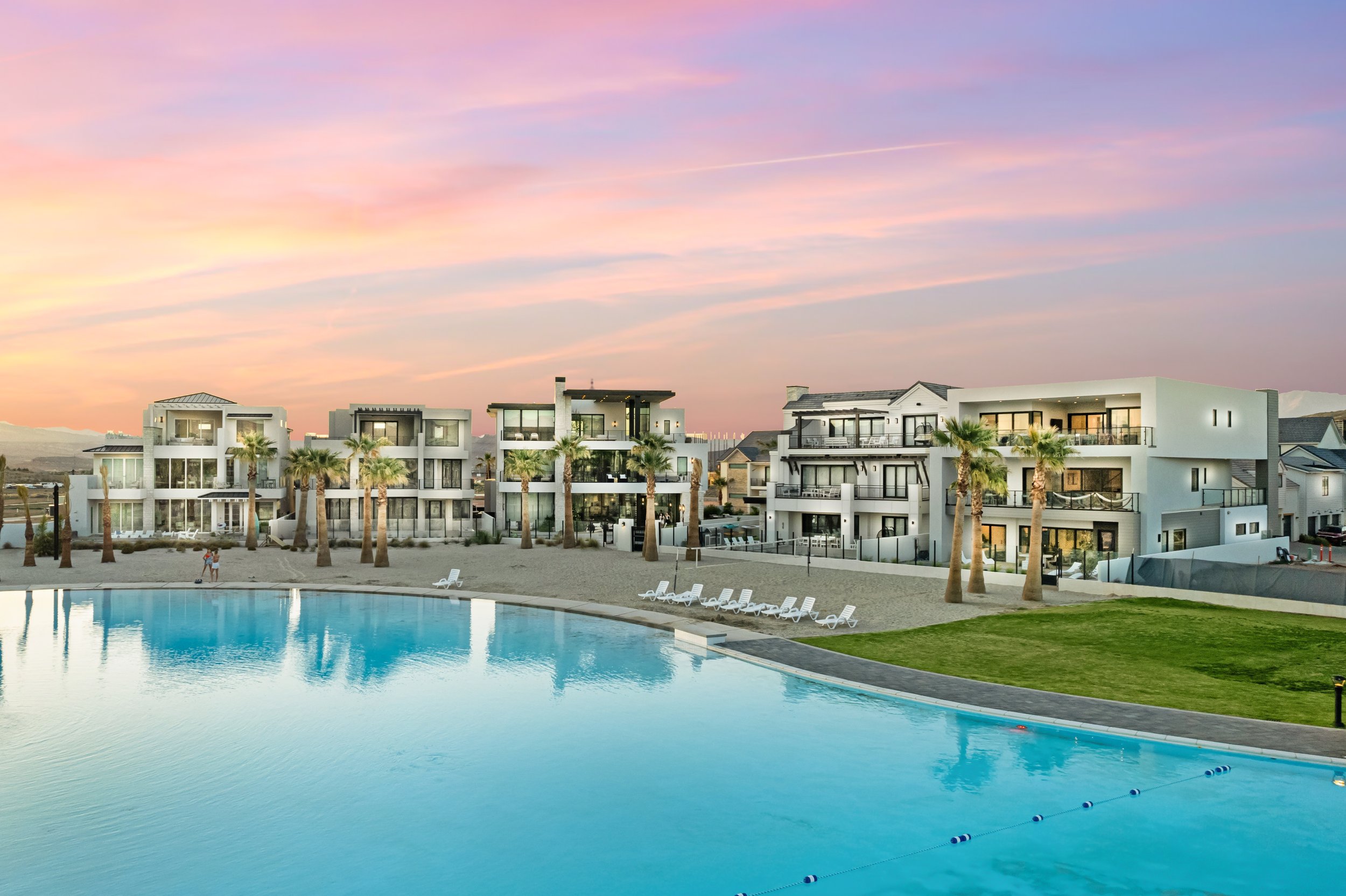 Luxury residential buildings with balconies and glass windows, a swimming pool in the foreground, beach with palm trees, and a colorful sunset sky.