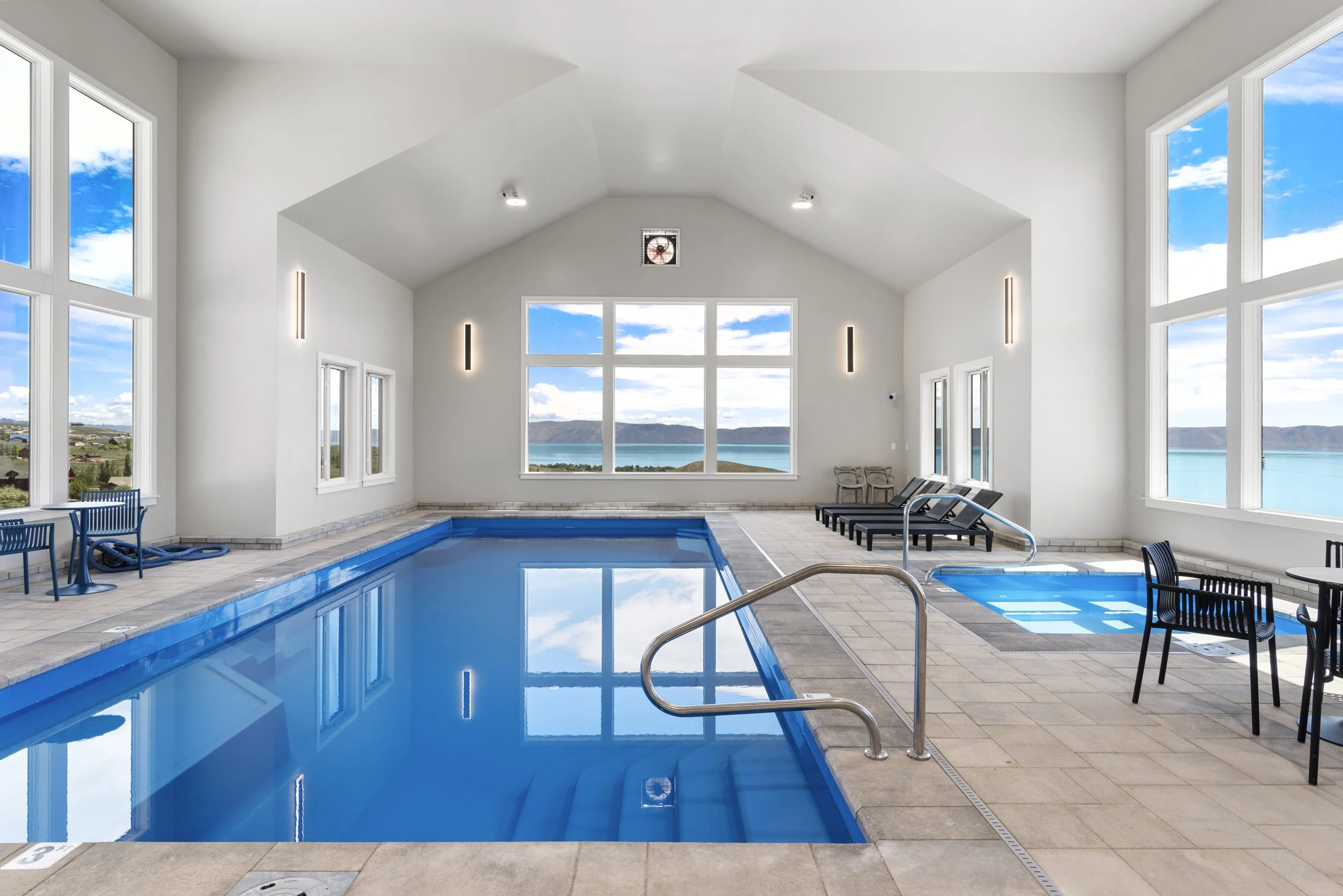 Indoor swimming pool with large windows overlooking a scenic view of water and hills, with lounge chairs and tables around the pool area, and a smaller hot tub.
