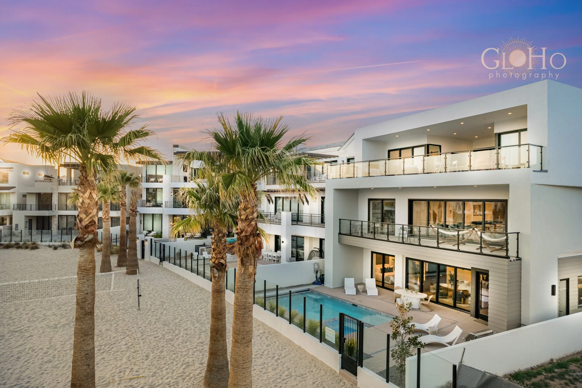 Exterior view of a modern beachfront luxury complex during sunset, with sand, palm trees, and a swimming pool.
