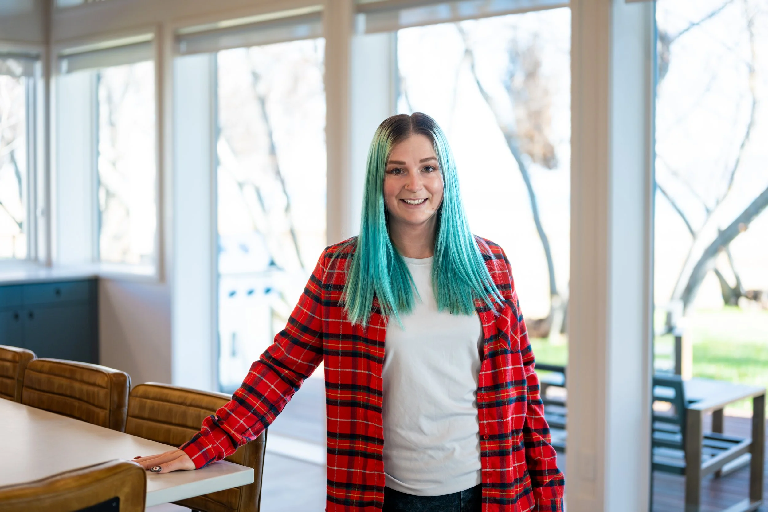 A woman with teal hair and a red plaid shirt standing in a bright indoor space with large windows and a view of trees outside.