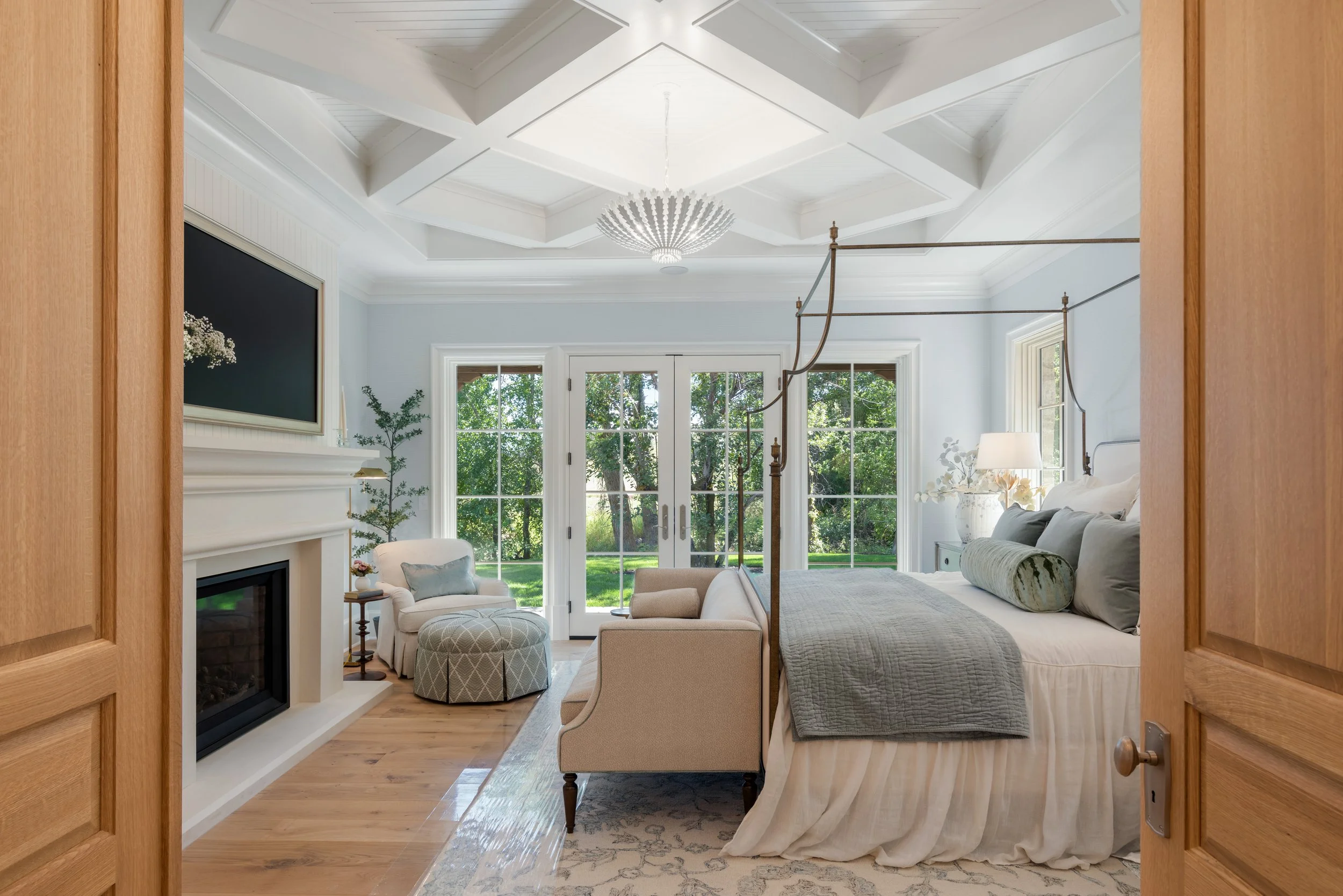 A bedroom with a four-poster bed, a beige armchair with a matching ottoman, a fireplace, large windows and glass doors overlooking a green yard, and a chandelier hanging from a coffered ceiling.