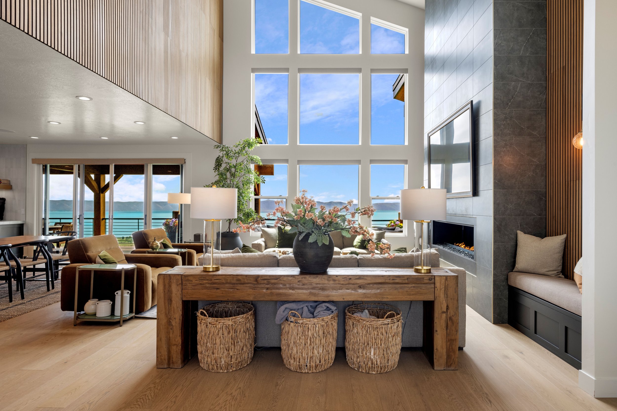 Living room with large windows showing blue sky and lake view, modern fireplace, TV, beige sofa, armchairs, lamps, and decorative baskets.