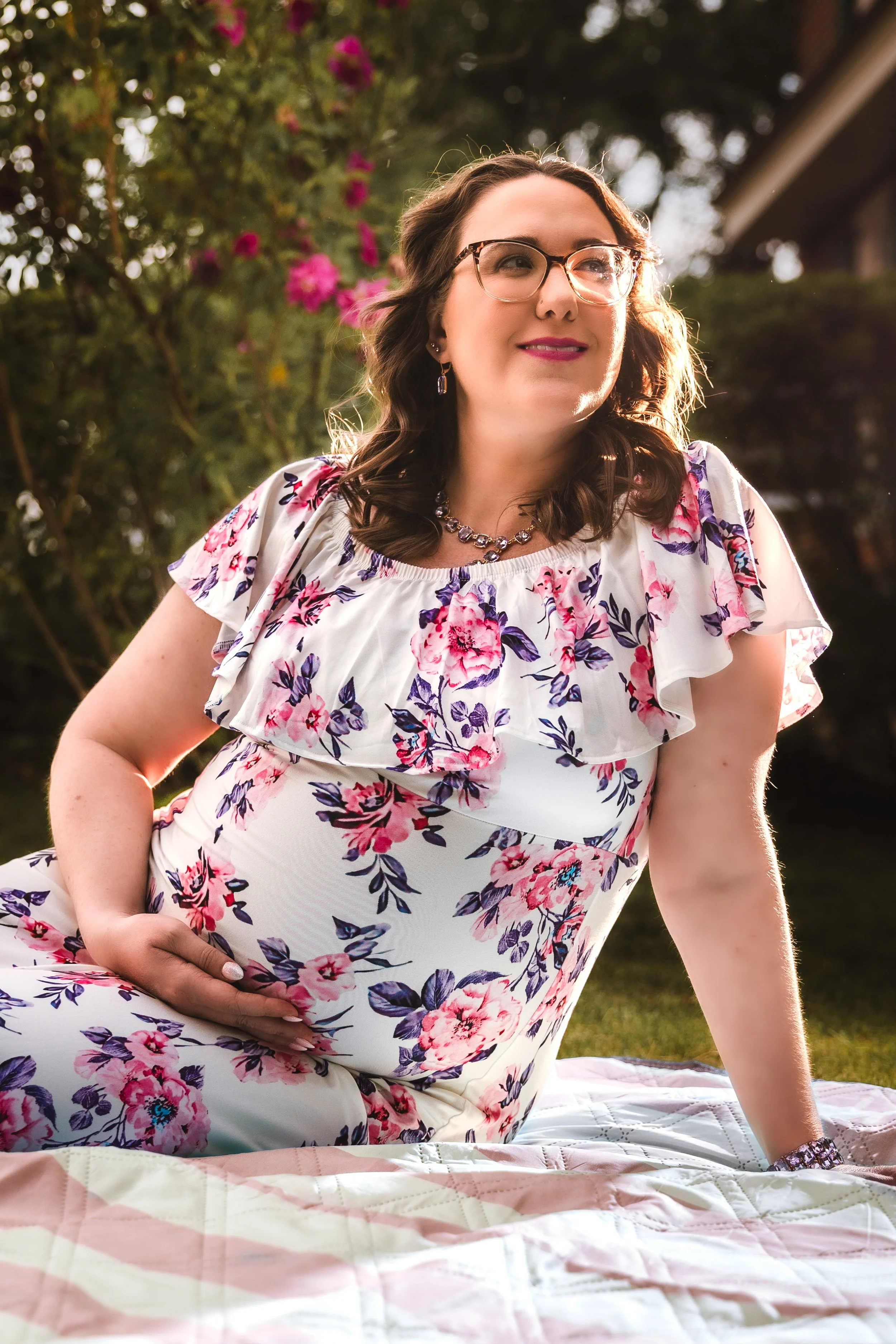 Pregnant woman sitting outdoors on a blanket, smiling and looking off to the side. She is wearing glasses, a floral dress, and jewelry, with flowers and greenery in the background.