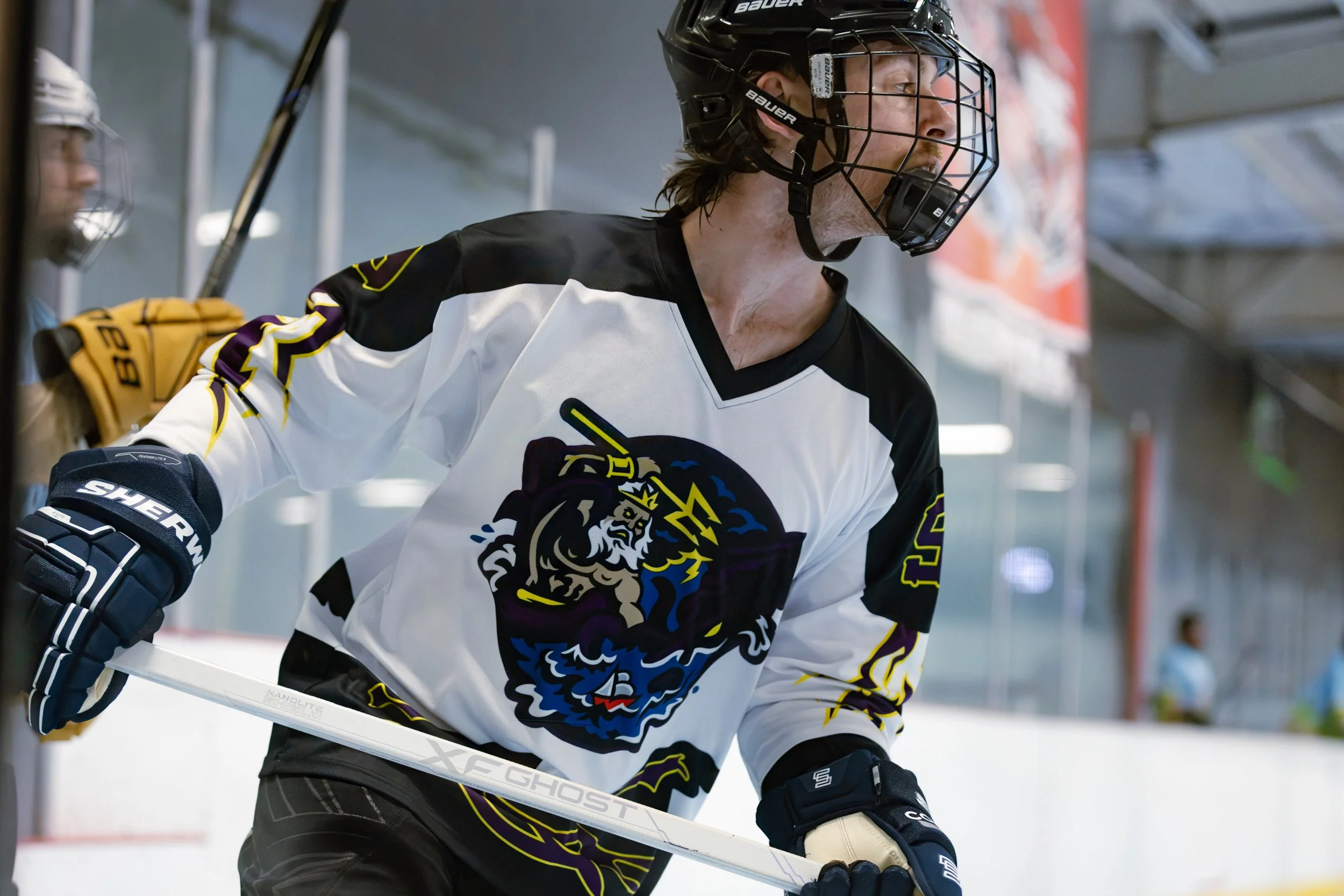 Hockey player in black and white jersey with a lion's head logo, wearing a black helmet and gloves, holding a white hockey stick on the ice rink.