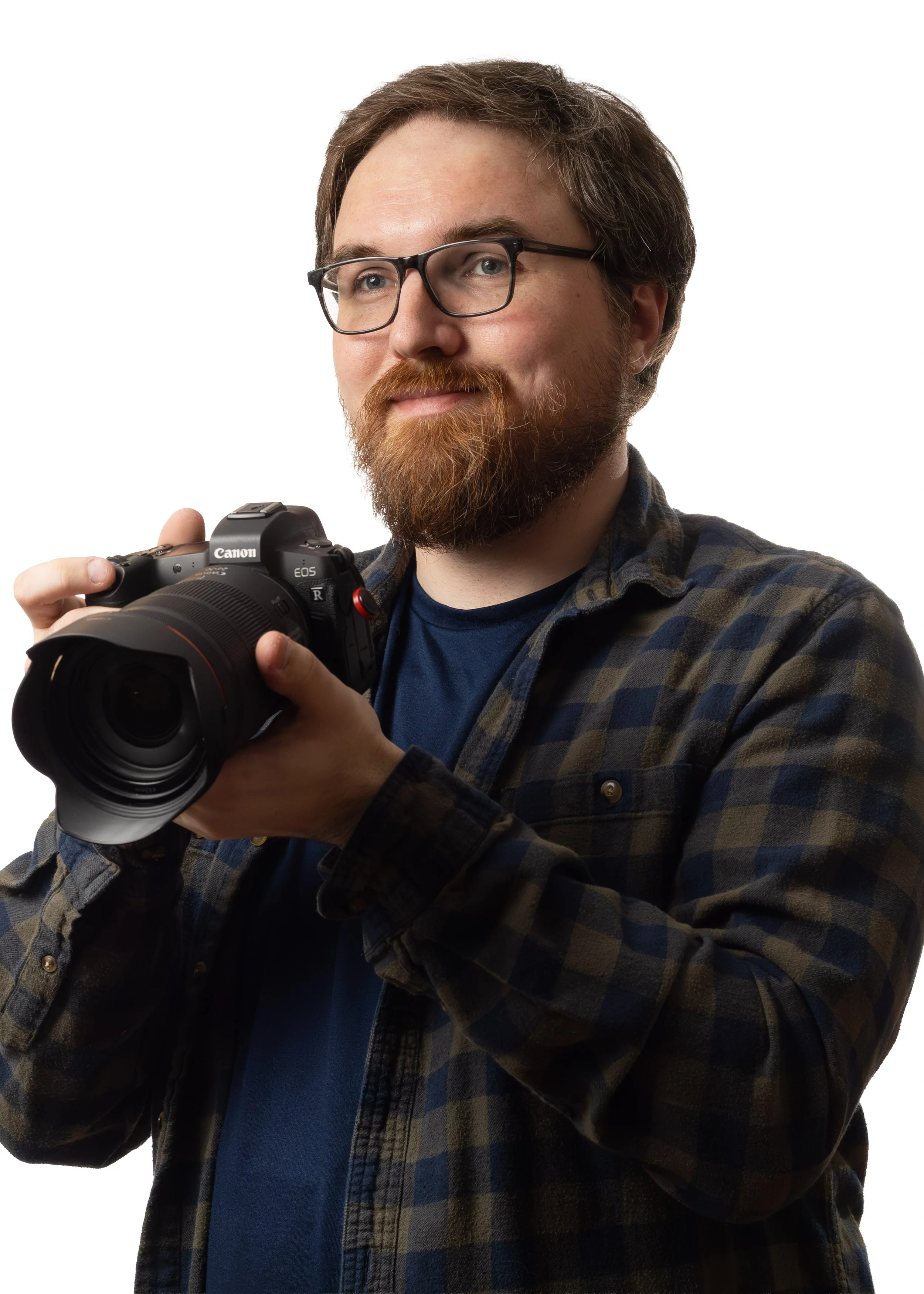 A man with glasses, a beard, and a mustache smiling while holding a Canon EOS camera, wearing a checkered shirt over a blue t-shirt, with a plain white background.