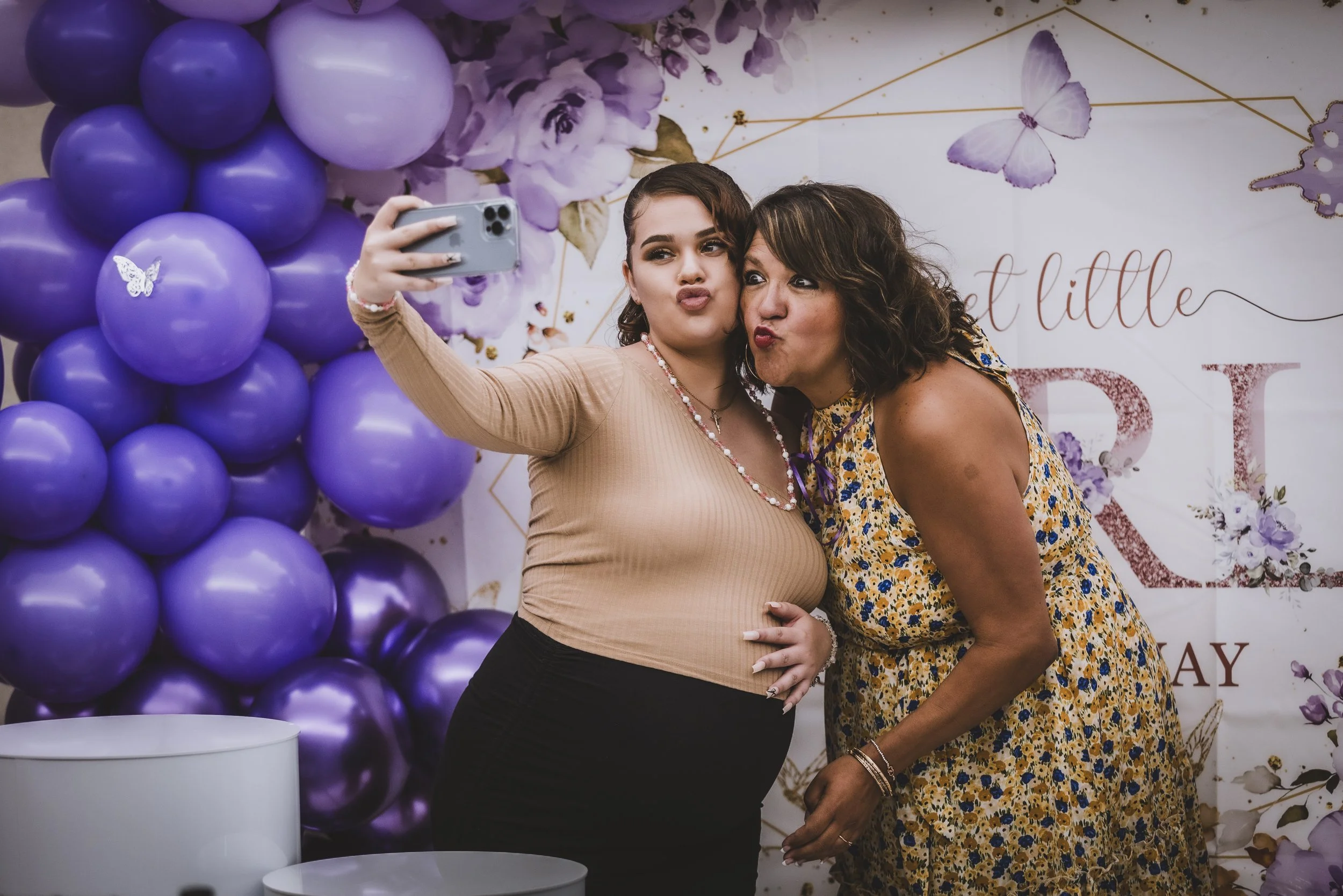 Two women taking a selfie at a celebration with purple balloons and floral decorations in the background.