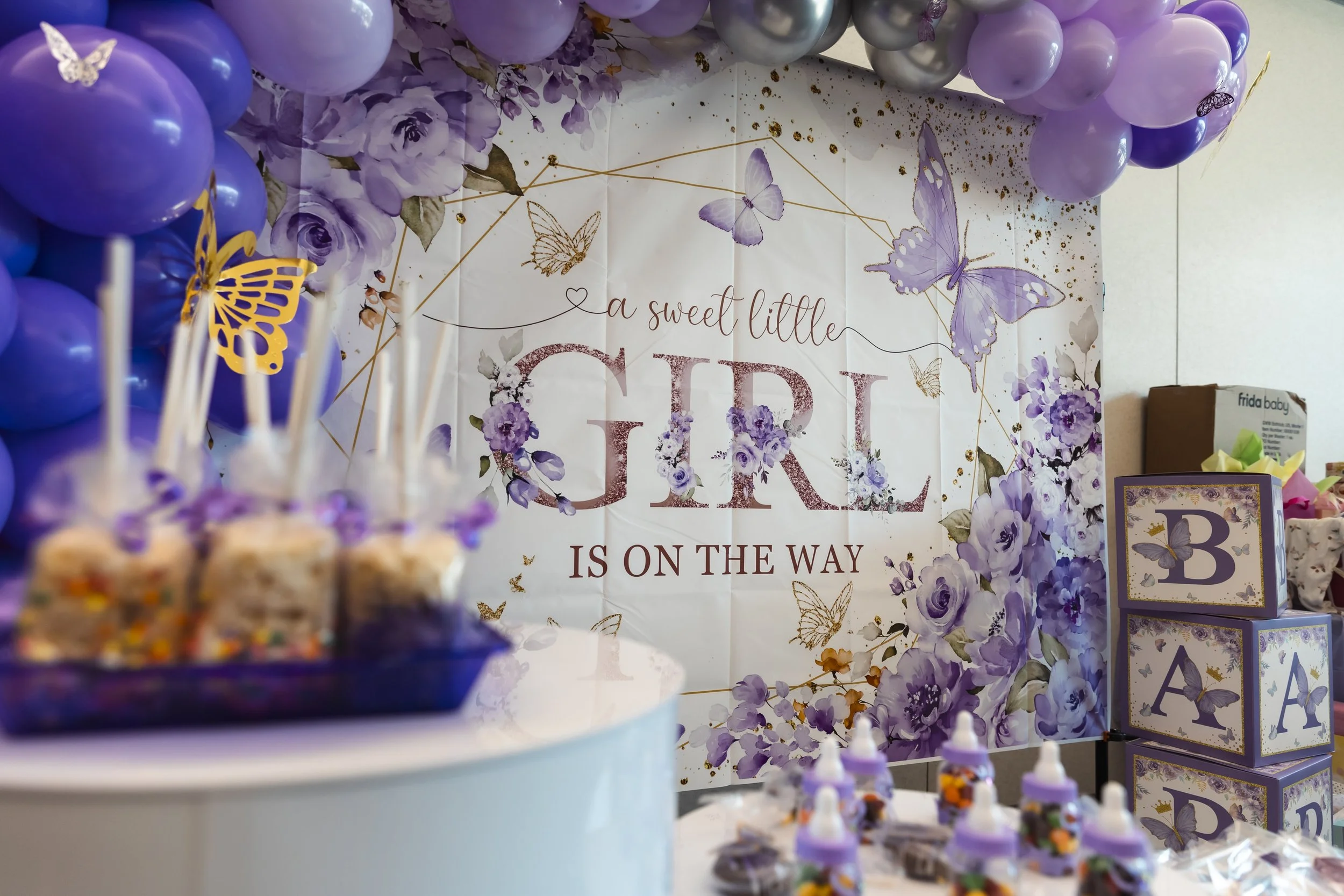 Decorative setup for a baby shower or birthday party with purple and lavender balloons, a floral backdrop with butterflies, and a sign that reads 'a sweet little girl is on the way'. There are also baby blocks and bottles on display.