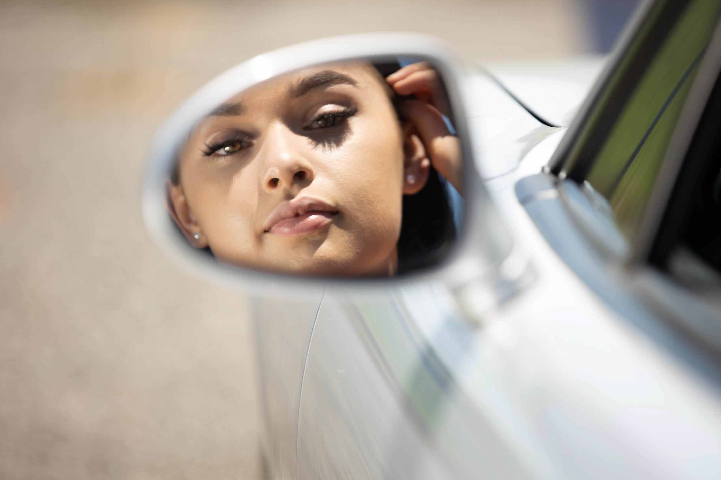 Close-up of a woman's face looking into a car's side mirror, with her hand behind her ear.