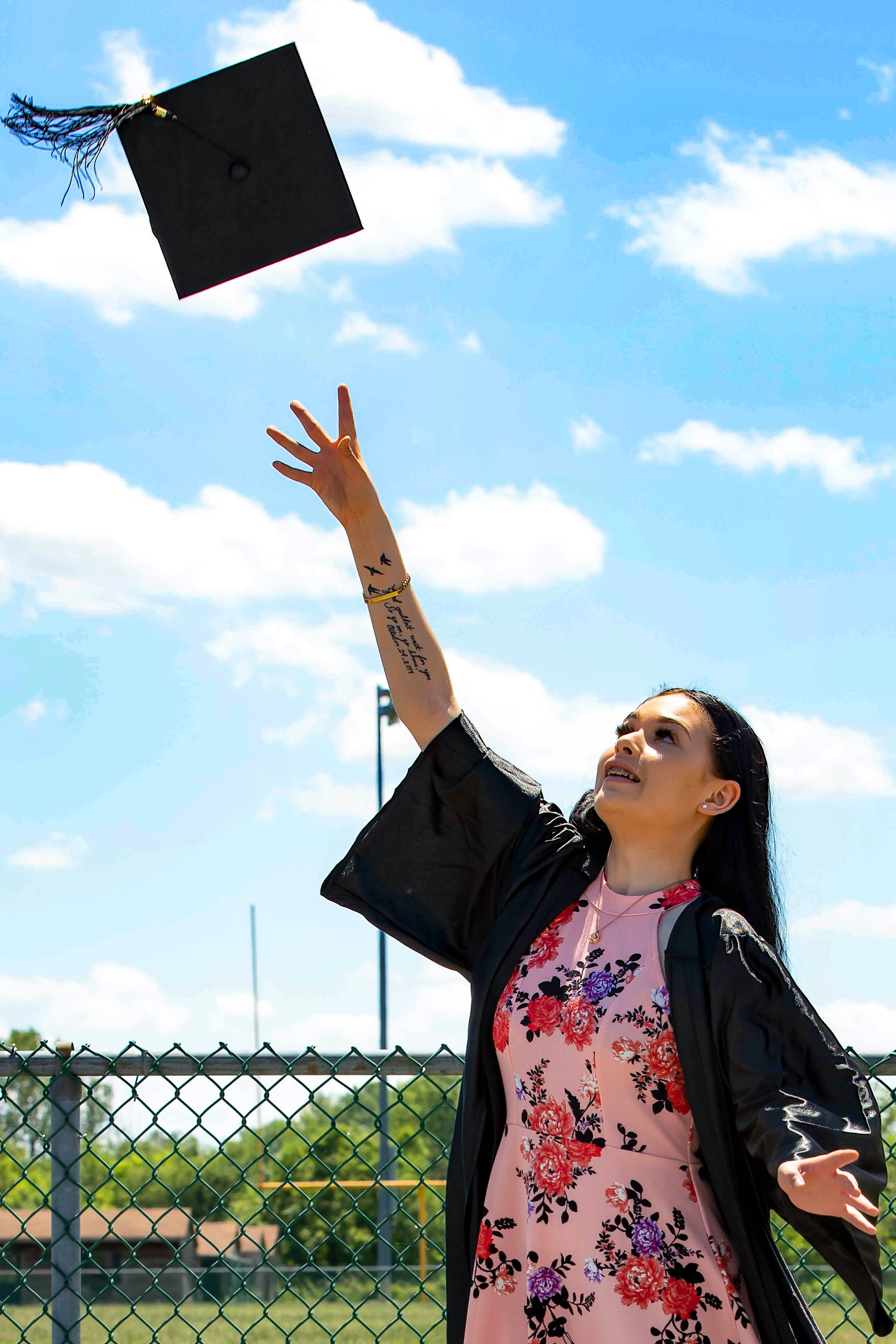 A young woman in a black graduation gown and floral dress throws her graduation cap into the air during a sunny outdoor graduation ceremony. Graduation photography by Digital Imageries Photography.