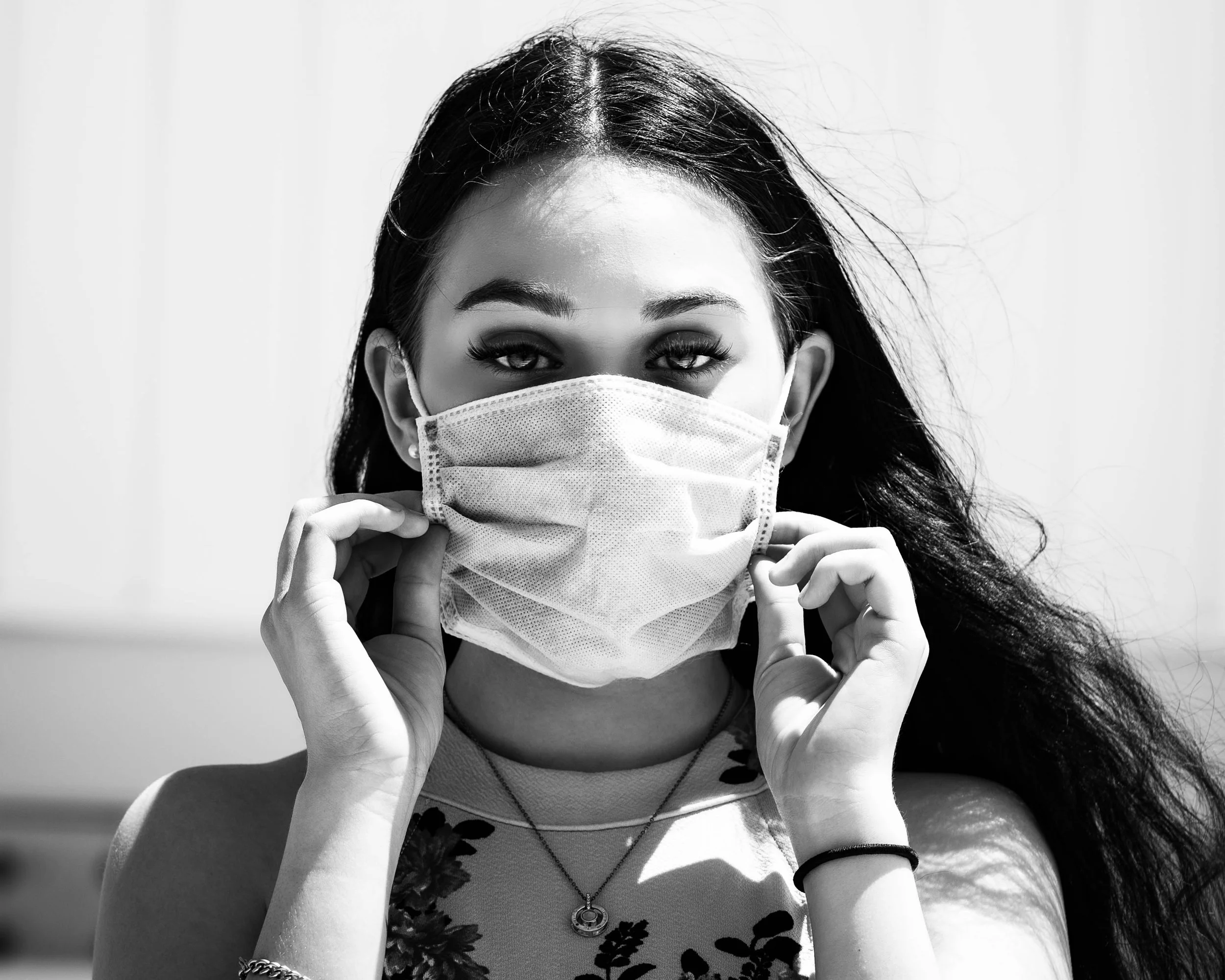 Black and white photo of a young woman with long hair, wearing a surgical mask, holding it with both hands, and looking directly at the camera.