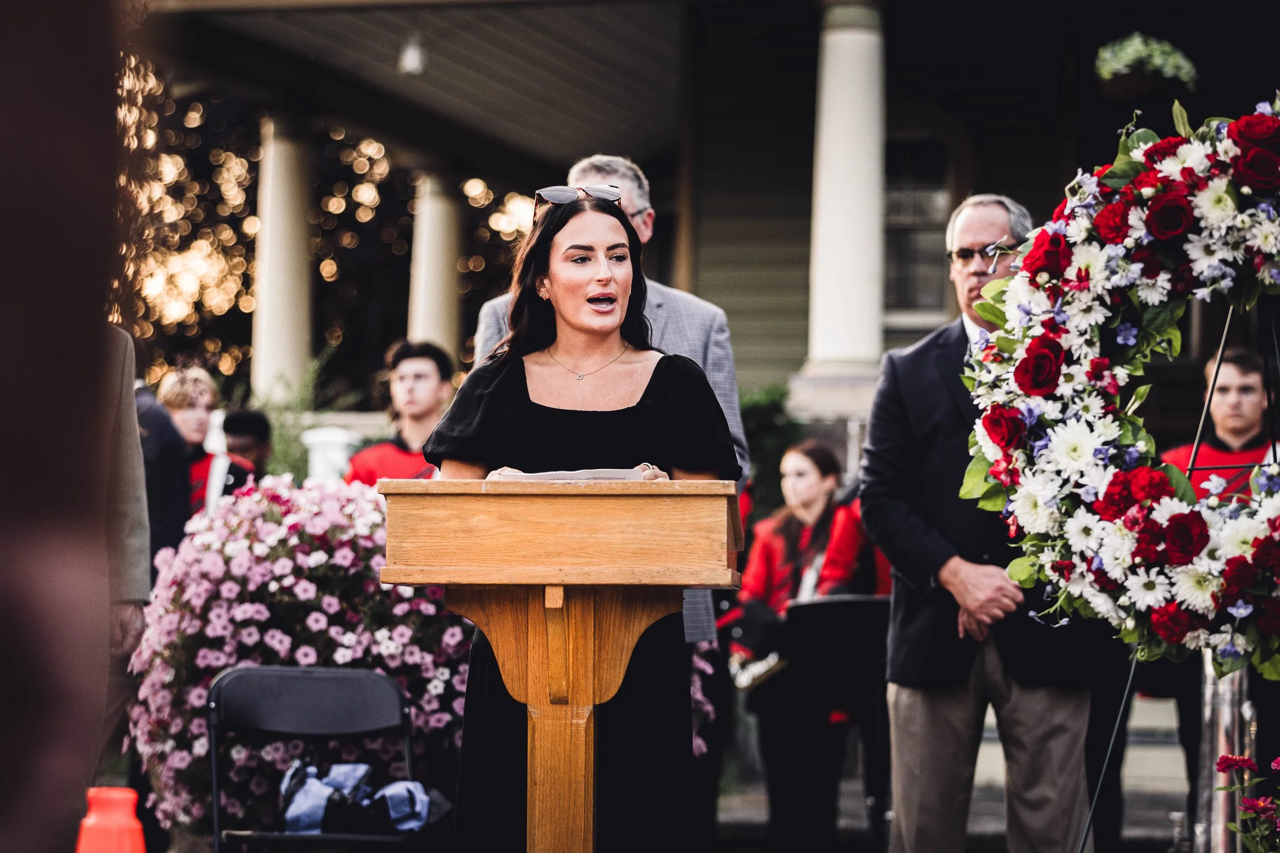 A woman speaking at a wooden podium during an outdoor event, with people standing and sitting behind her, including a man in glasses and a suit, and framed by large floral arrangements.