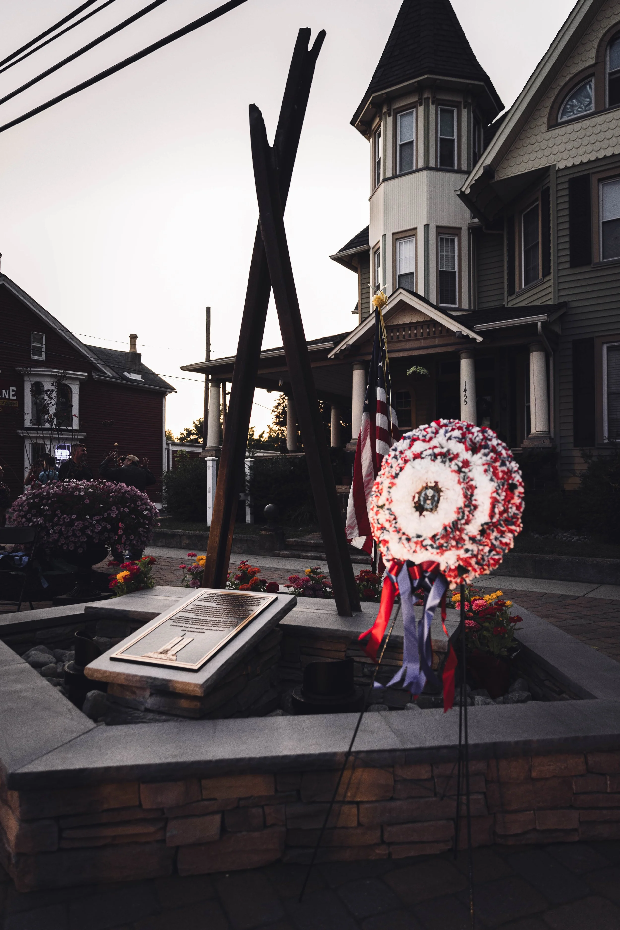 Memorial site with a cross and a wreath honor fallen soldiers, with an American flag and flowers in the background, in front of a Victorian-style house during sunset.
