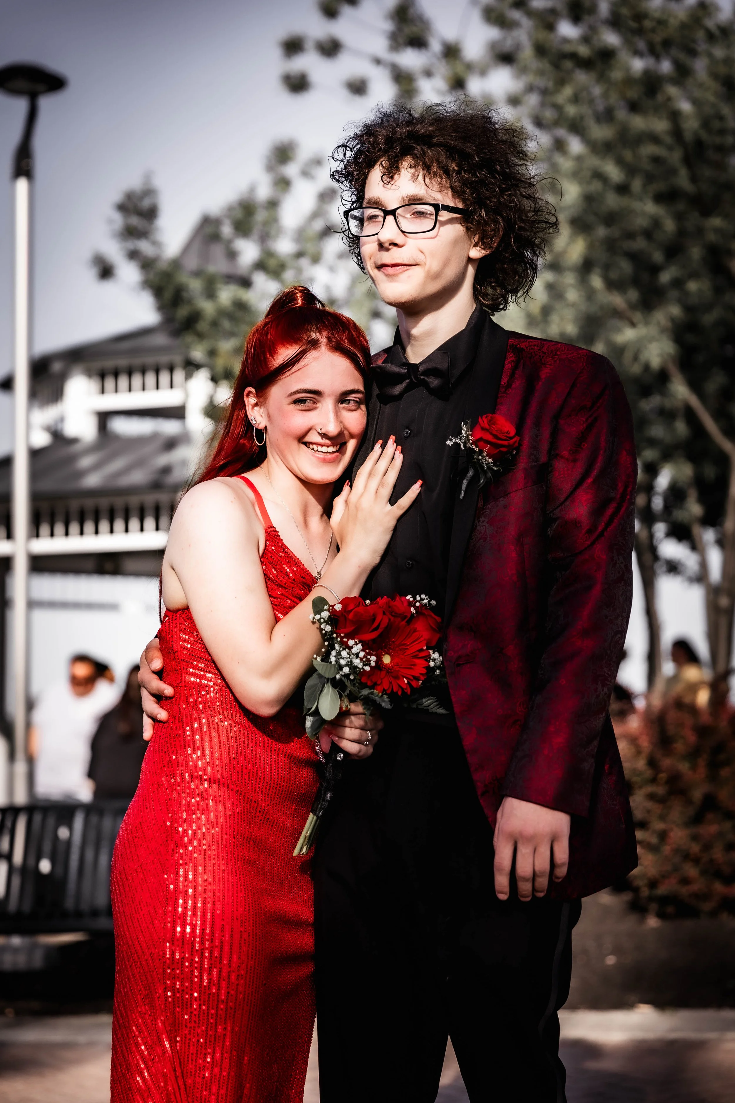 A young woman with red hair in a red dress and a young man with curly hair in a dark red tuxedo jacket and black shirt pose together outdoors, smiling and embracing. The woman holds a bouquet of red flowers, and the man has a red flower in his lapel.