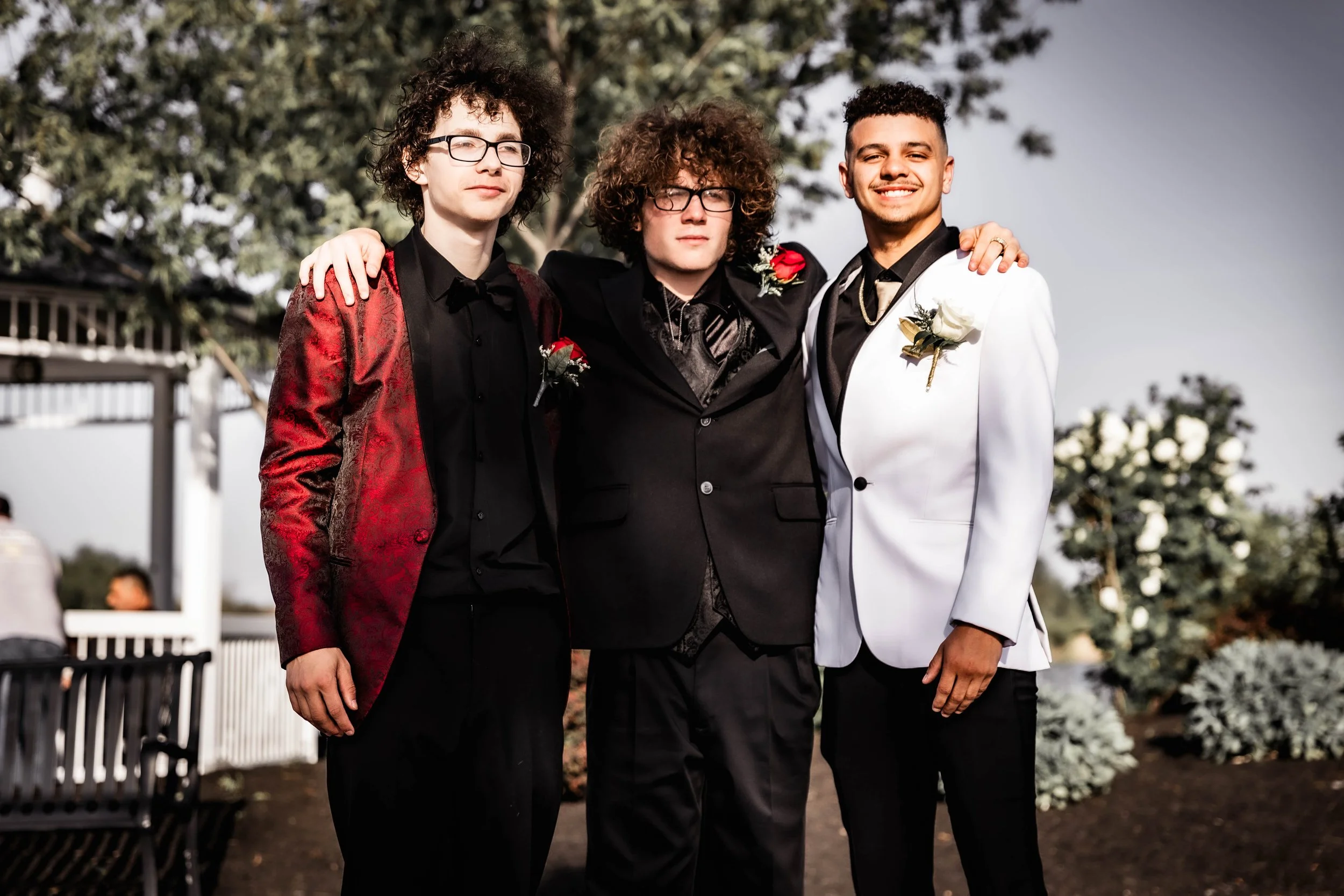 Three young men dressed in tuxedos, posing outdoors with arms around each other, smiling and looking at the camera.