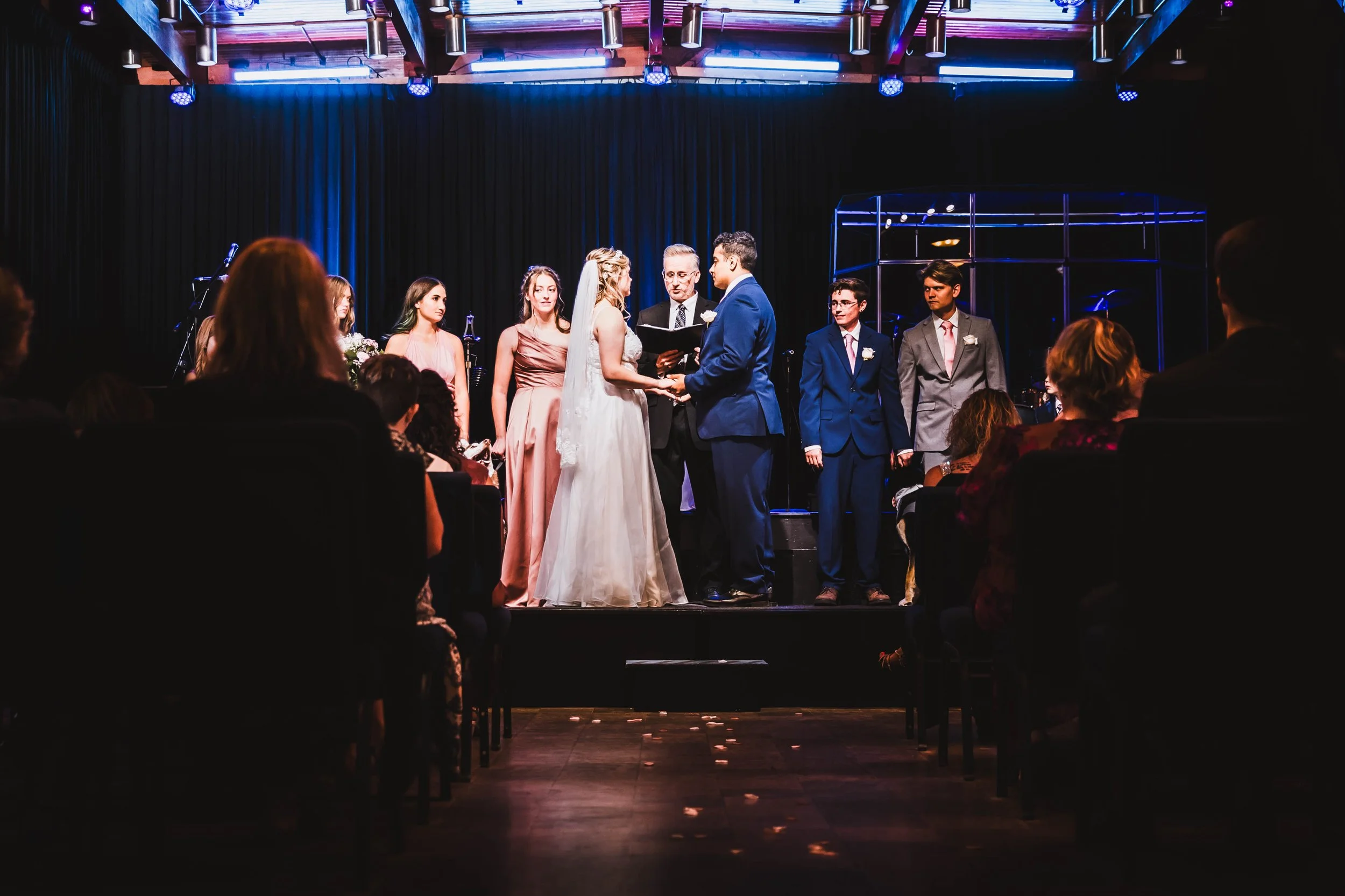 A wedding ceremony with a bride and groom exchanging vows on stage with an officiant, surrounded by bridesmaids and groomsmen, inside a dimly lit venue with purple and blue lighting.