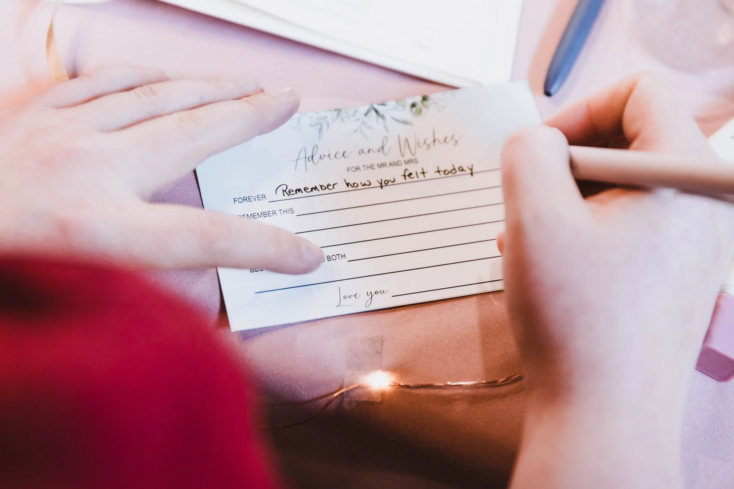 Close-up of a person writing on a card with a pen, with a pink tablecloth and string lights in the background.