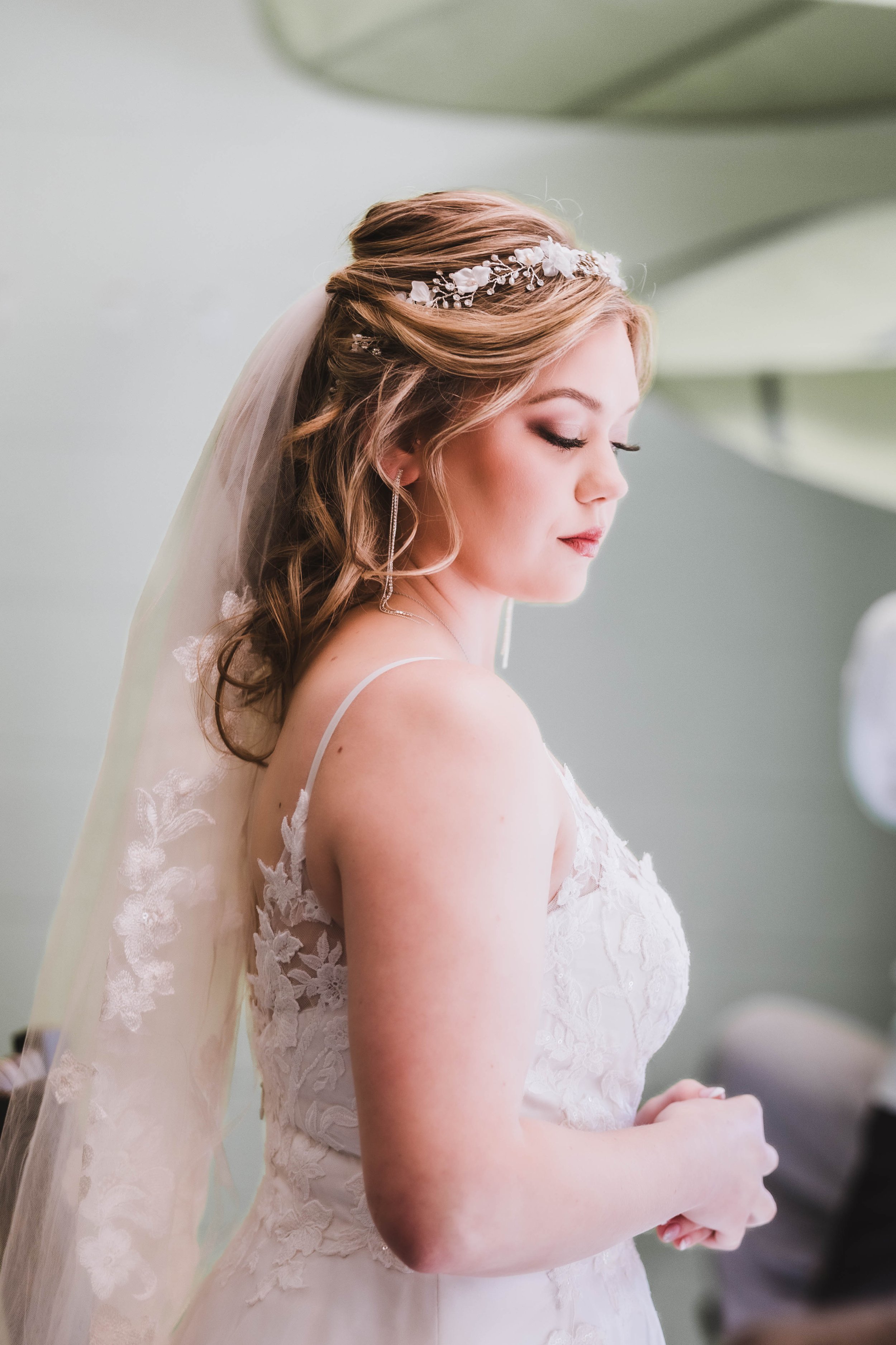 A bride with long, wavy hair and a delicate floral headband, wearing a lace wedding dress, standing with her eyes closed and hands clasped.