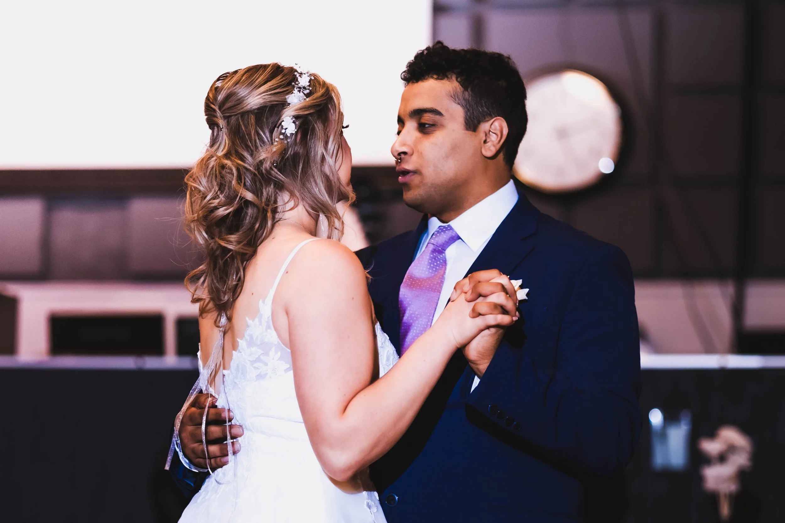 A couple dancing at their wedding, with the bride in a white dress and the groom in a navy suit, holding hands closely and looking into each other's eyes.