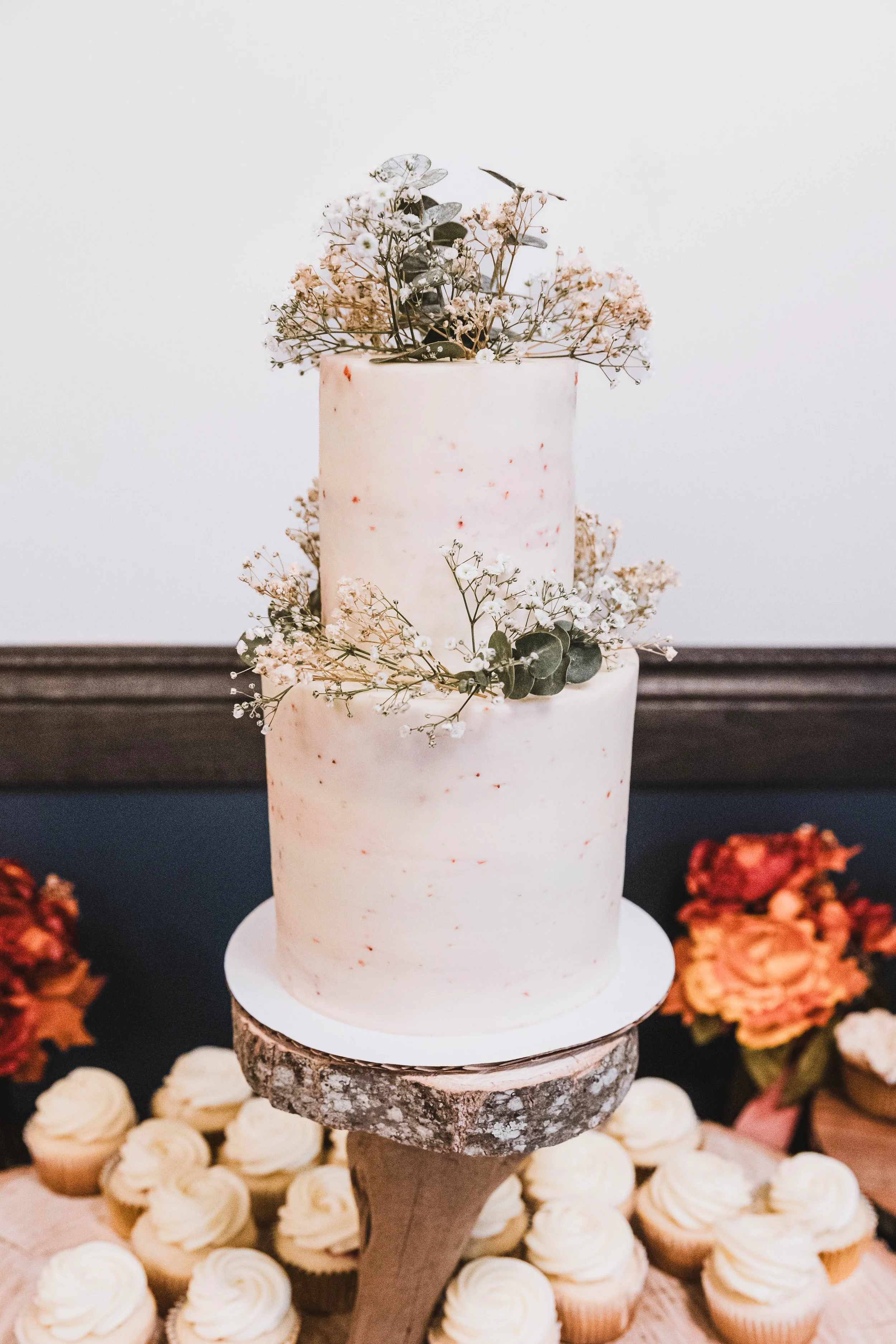 A two-tiered wedding or celebration cake with white frosting, decorated with small sprigs of baby's breath and eucalyptus, placed on a wooden cake stand. In the background are cupcakes with swirled frosting and some flowers.