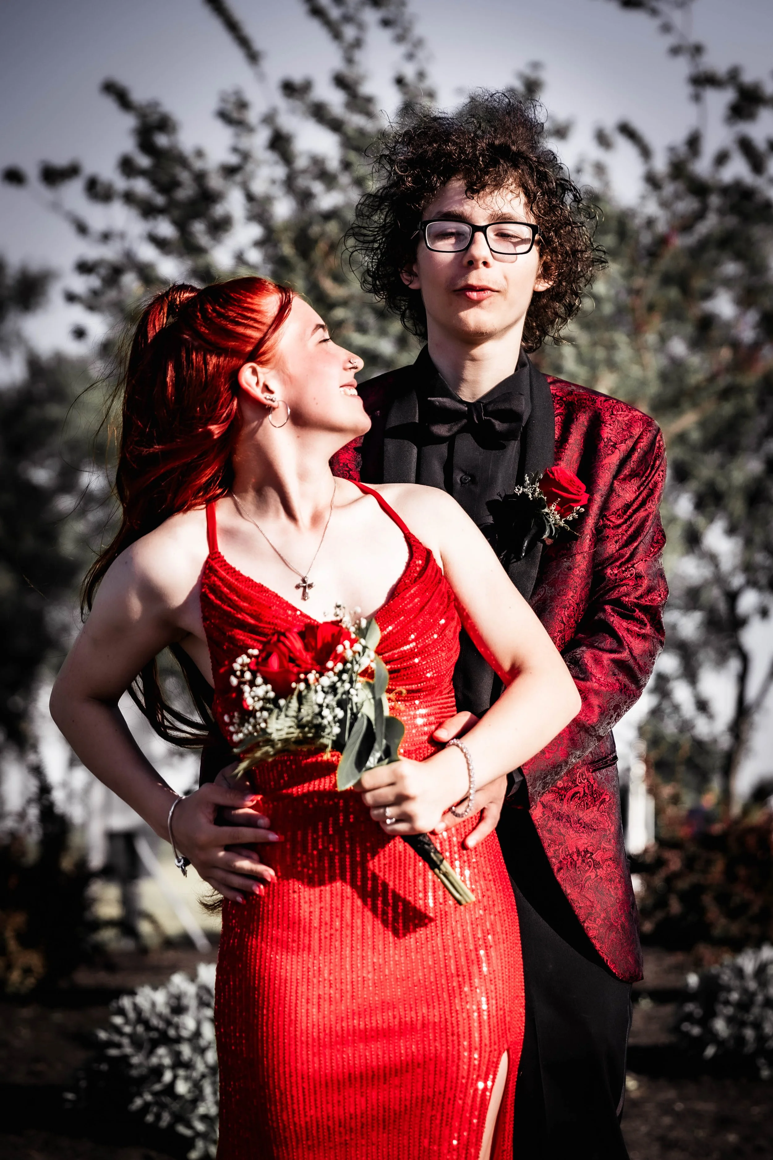 A couple in formal attire, the woman in a red dress holding a bouquet and the man in a matching red tuxedo jacket with black shirt and bow tie, standing outdoors.