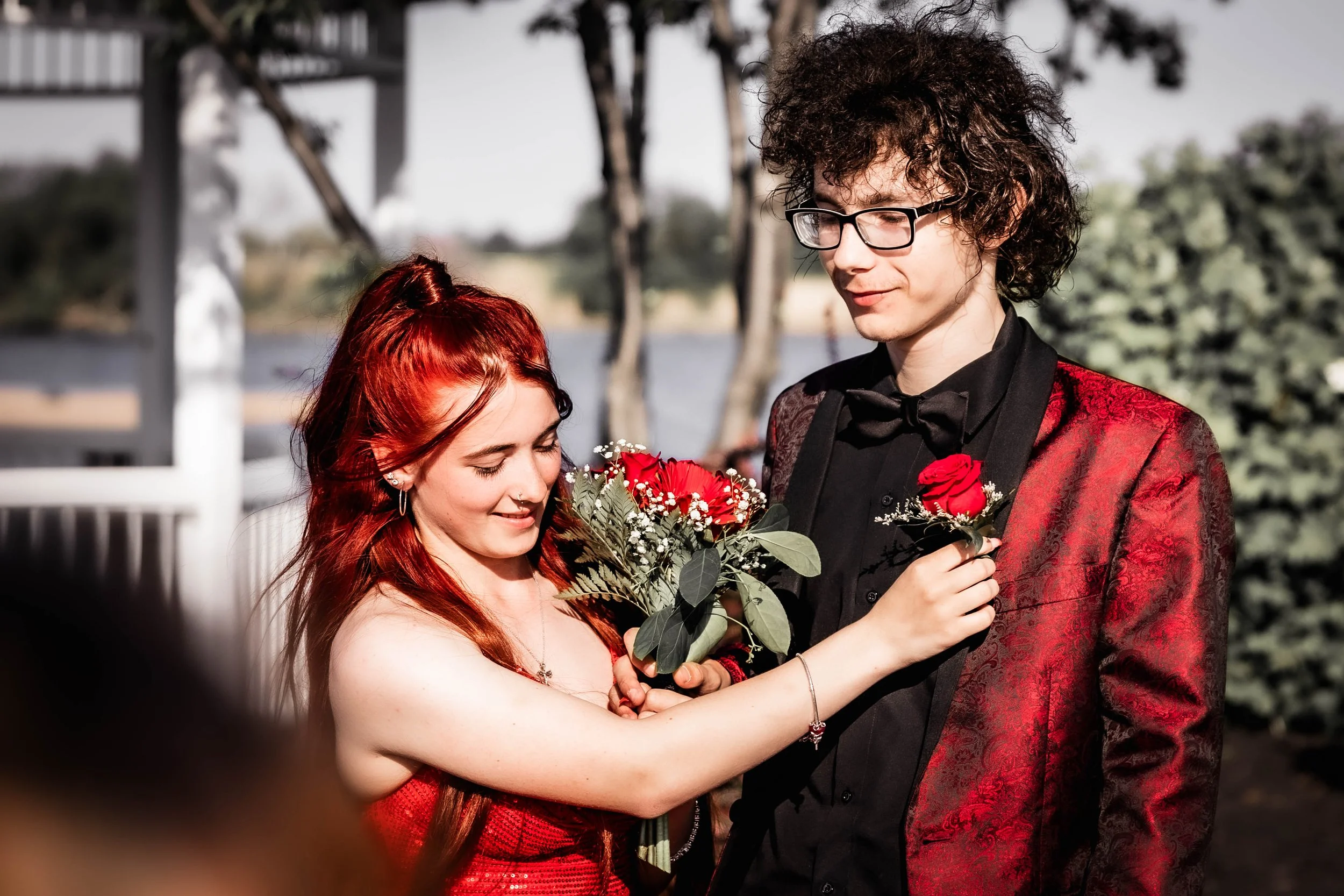 Young woman with red hair handing a red rose to a young man with dark curly hair and glasses, who is dressed in a red and black suit, outdoors with trees and a body of water in the background. Prom photography in South Jersey by Digital Imageries Pho