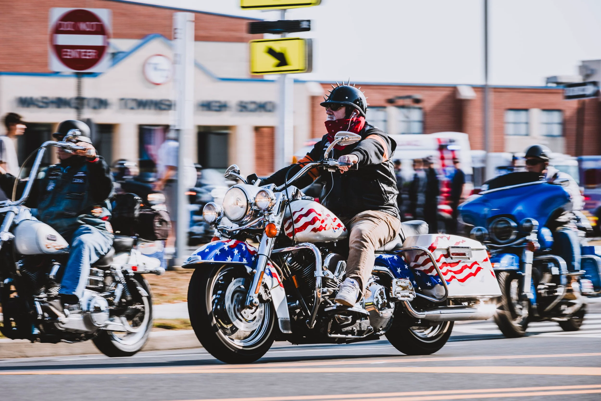 Motorcyclists riding on a city street with a patriotic American flag design on their bikes, wearing helmets and jackets.