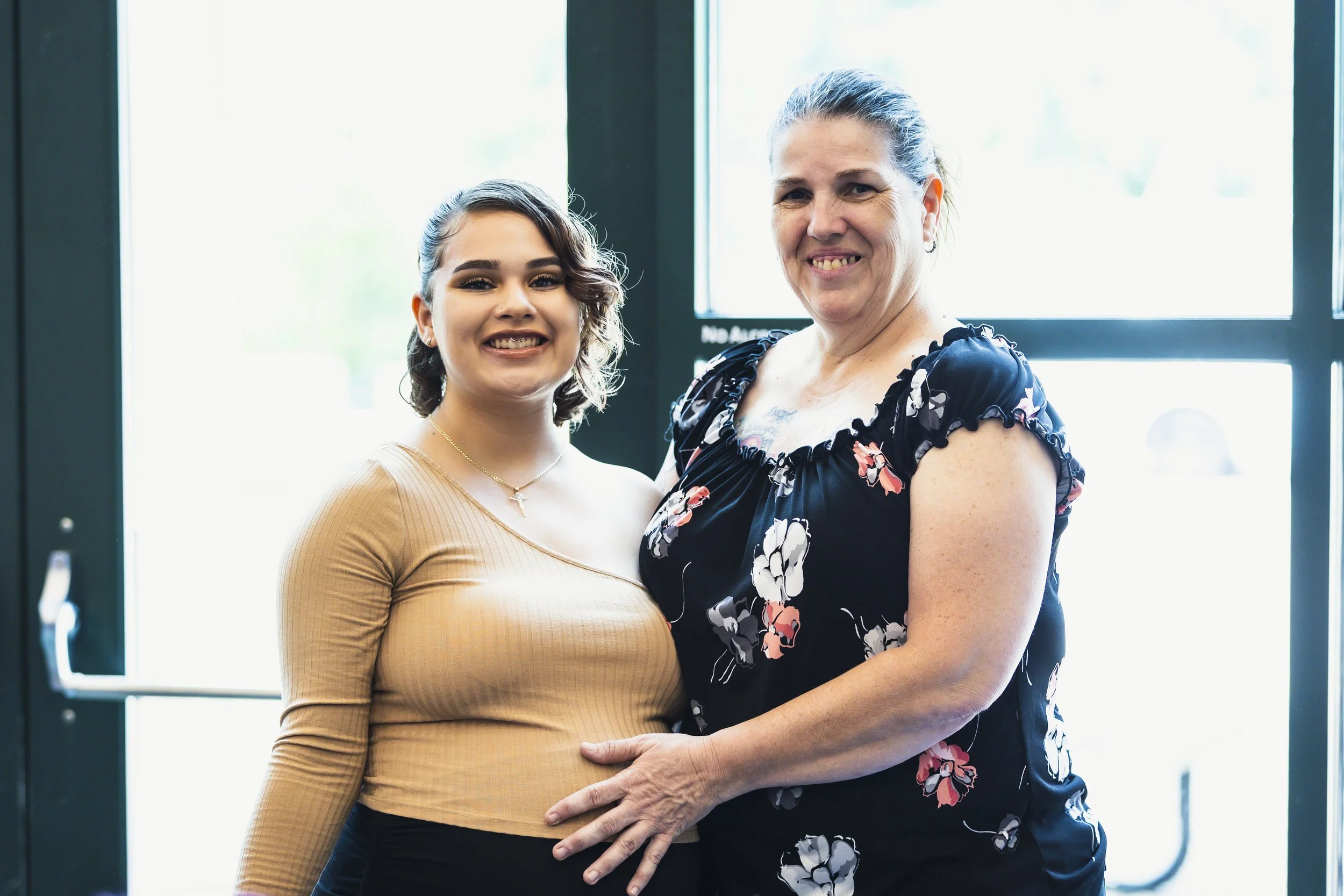 A young woman and an older woman standing together indoors by a glass door, smiling at the camera. The younger woman is wearing a beige shirt and a necklace, and the older woman is wearing a black floral dress and has her hand on the younger woman's 