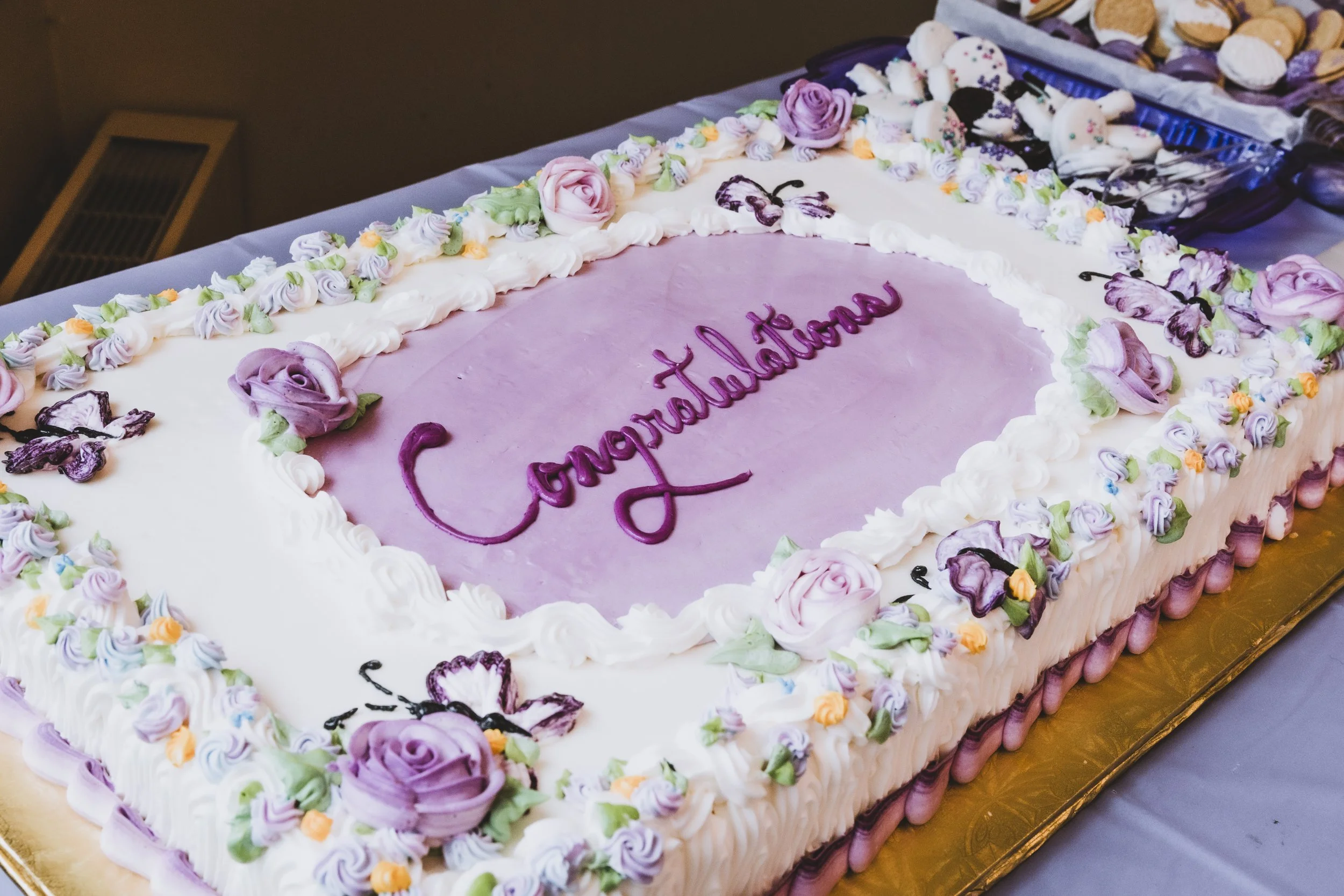 Large rectangular cake with white and purple icing, decorated with purple and pink roses, and the word 'Congratulation' written in purple icing in the center.