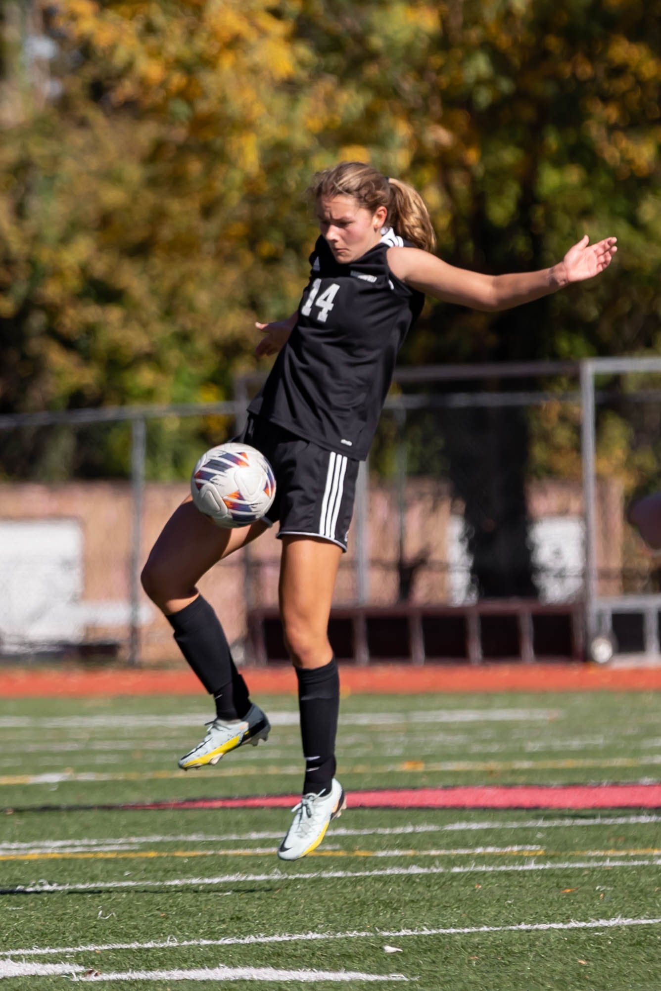 A soccer player in a dark uniform showing impressive athletic skill by controlling a ball in mid-air with her knee during a sunny afternoon match.
