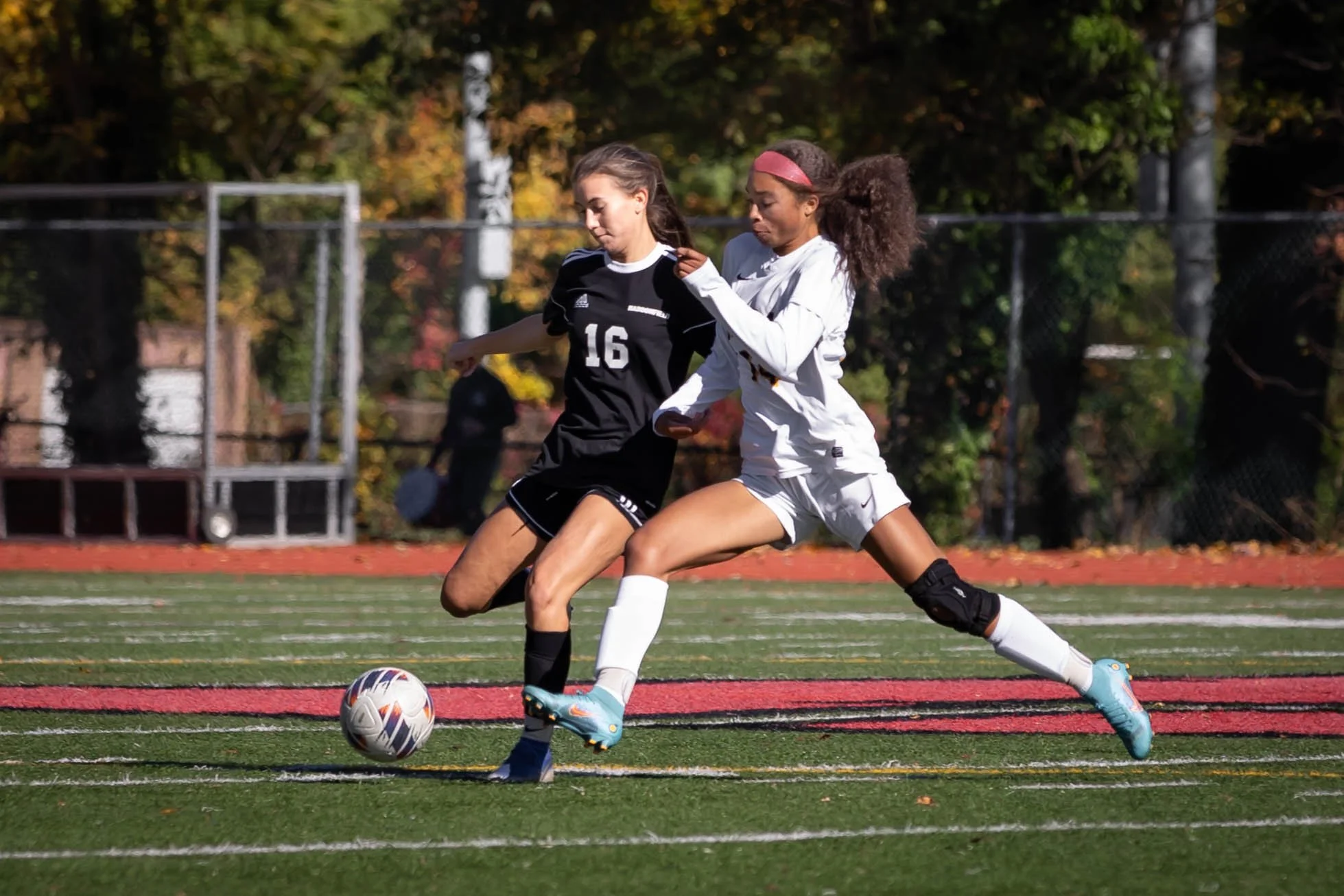 Two players from opposing teams in a fierce one-on-one battle for the soccer ball near the sideline on a bright turf field.